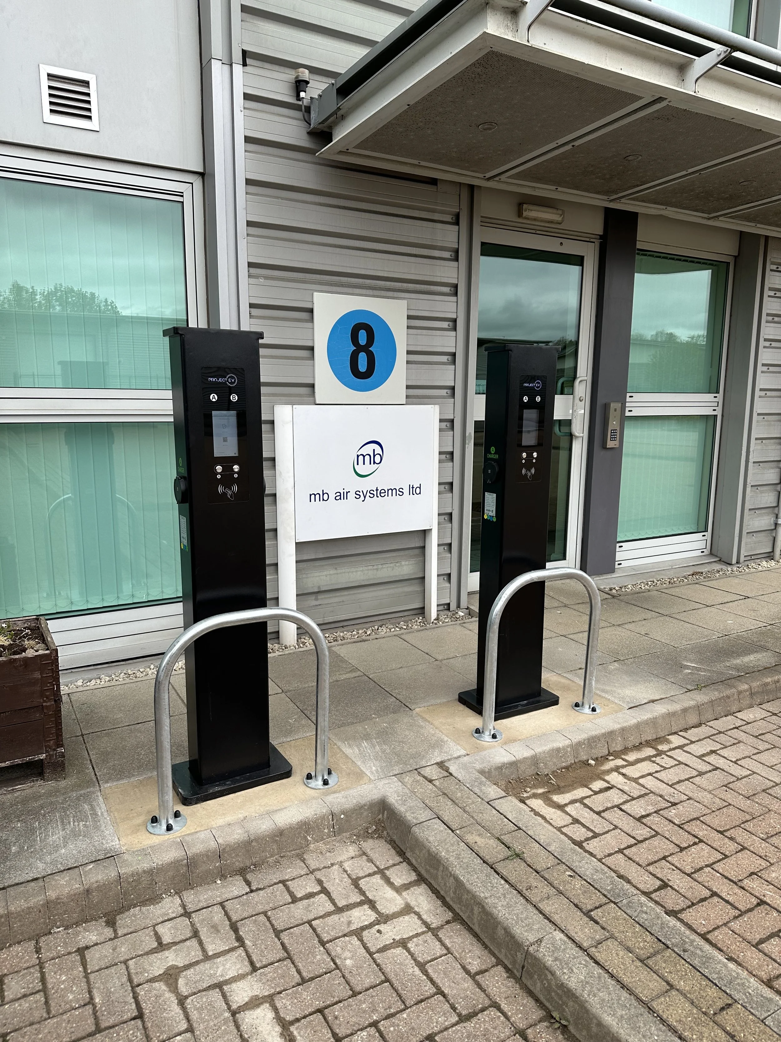 Outside of a building with two charging stations for electric vehicles, a sign with the number 8, and a company sign that reads 'mb air systems ltd'. The charging stations have screens and contactless payment options, and are protected by metal barriers.