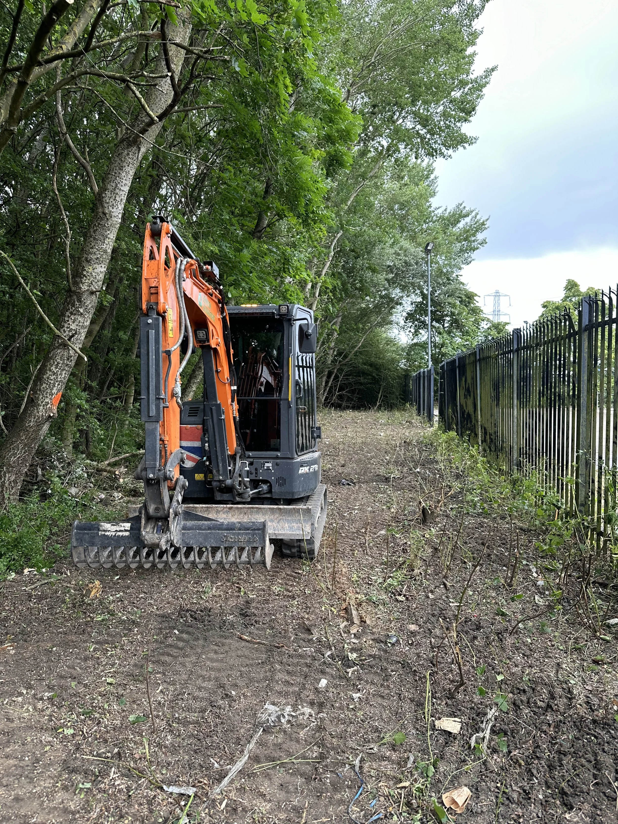 A small orange and black excavator working on a dirt path beside a black metal fence, surrounded by trees and greenery.