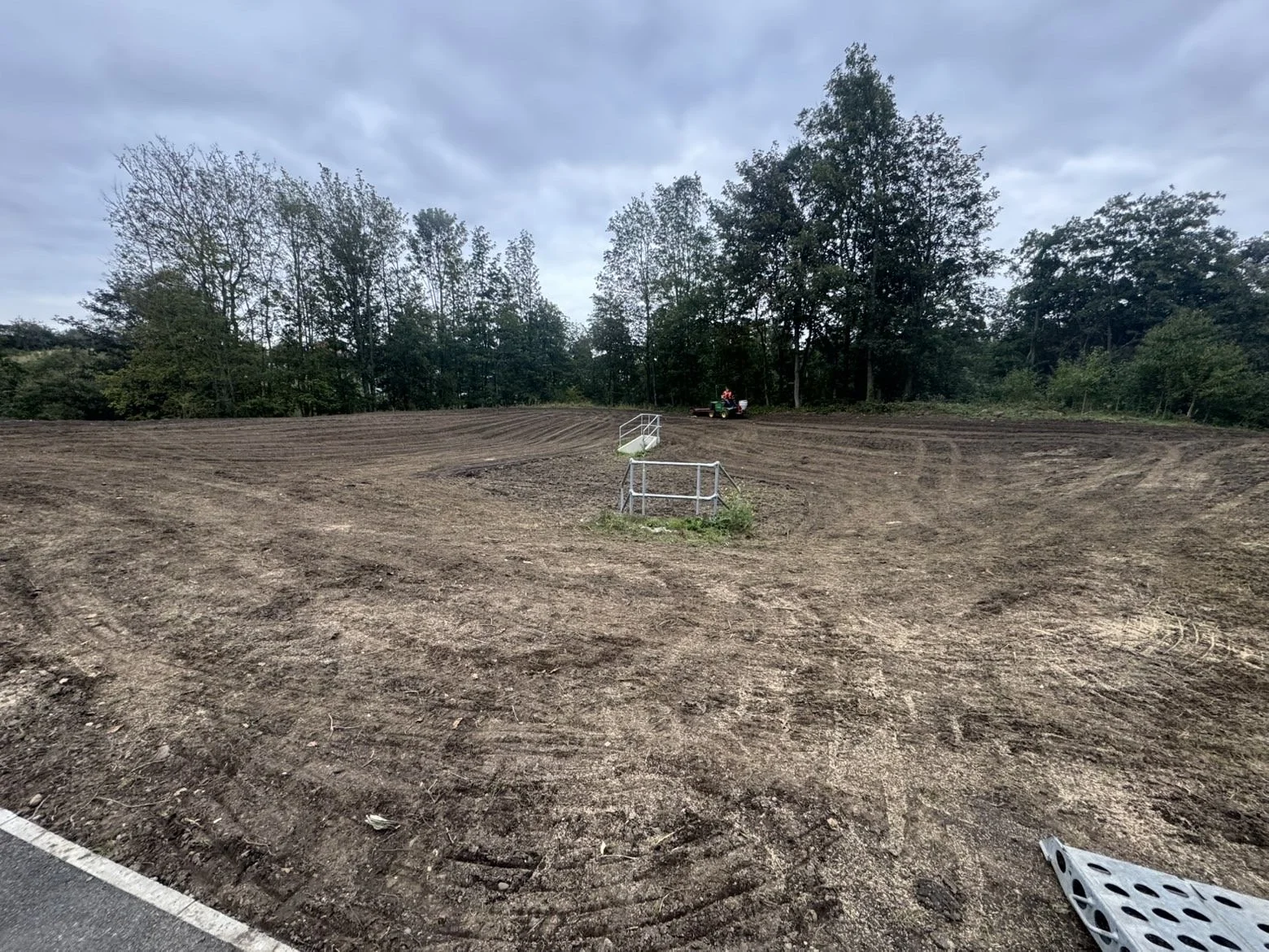 A cleared, inclined dirt field with a small metal fence in the foreground, trees in the background, overcast sky, and a riding mower with a person on it near the top of the hill.