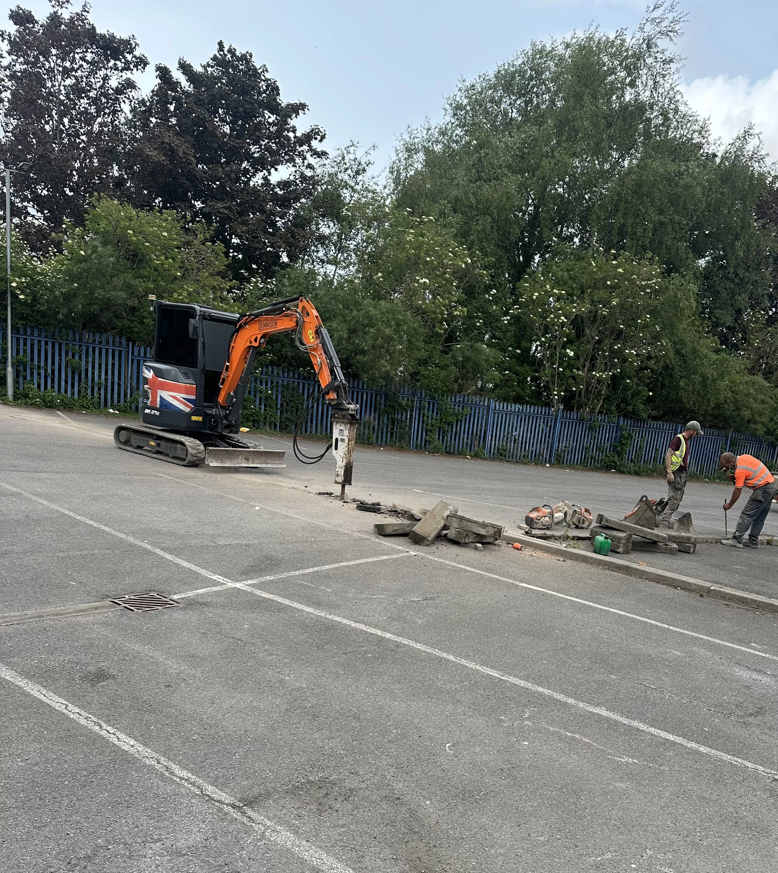 Construction workers and a small excavator working on a sidewalk next to a parking lot, with trees and a blue fence in the background.