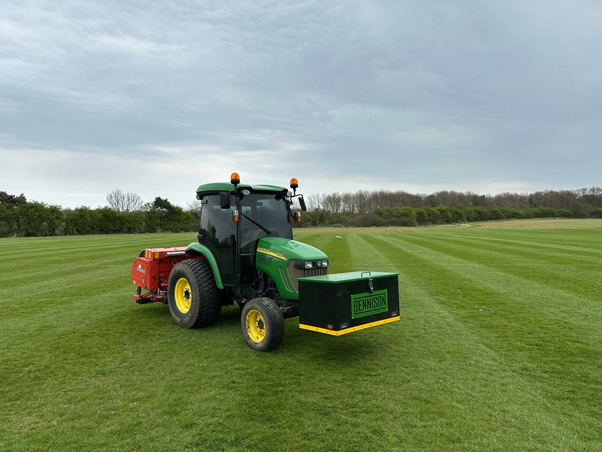 Green John Deere tractor on a well-manicured grassy field under a cloudy sky, with trees in the background.