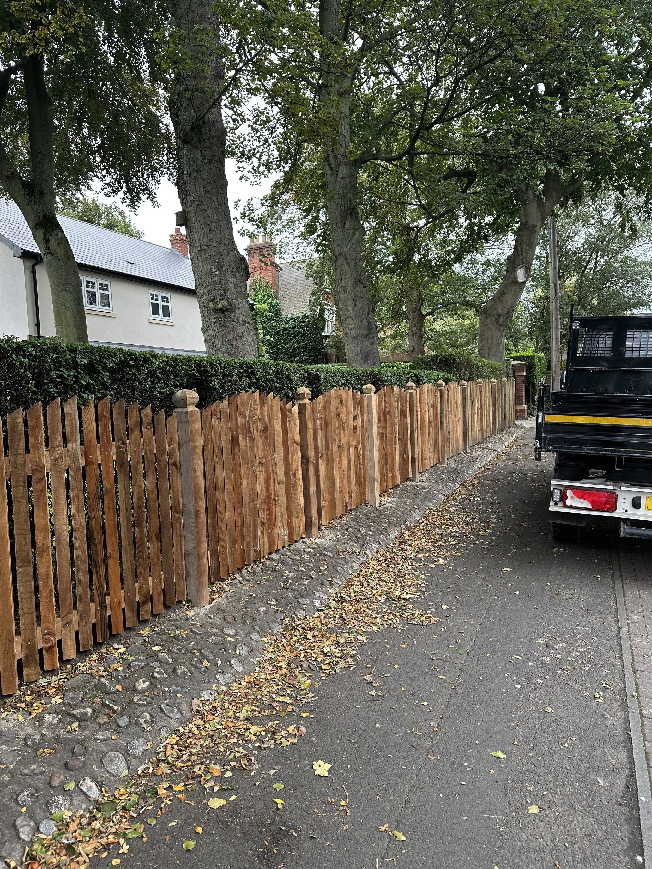A wooden picket fence along a sidewalk with fallen leaves, large trees in the background, and part of a black truck parked on the street.