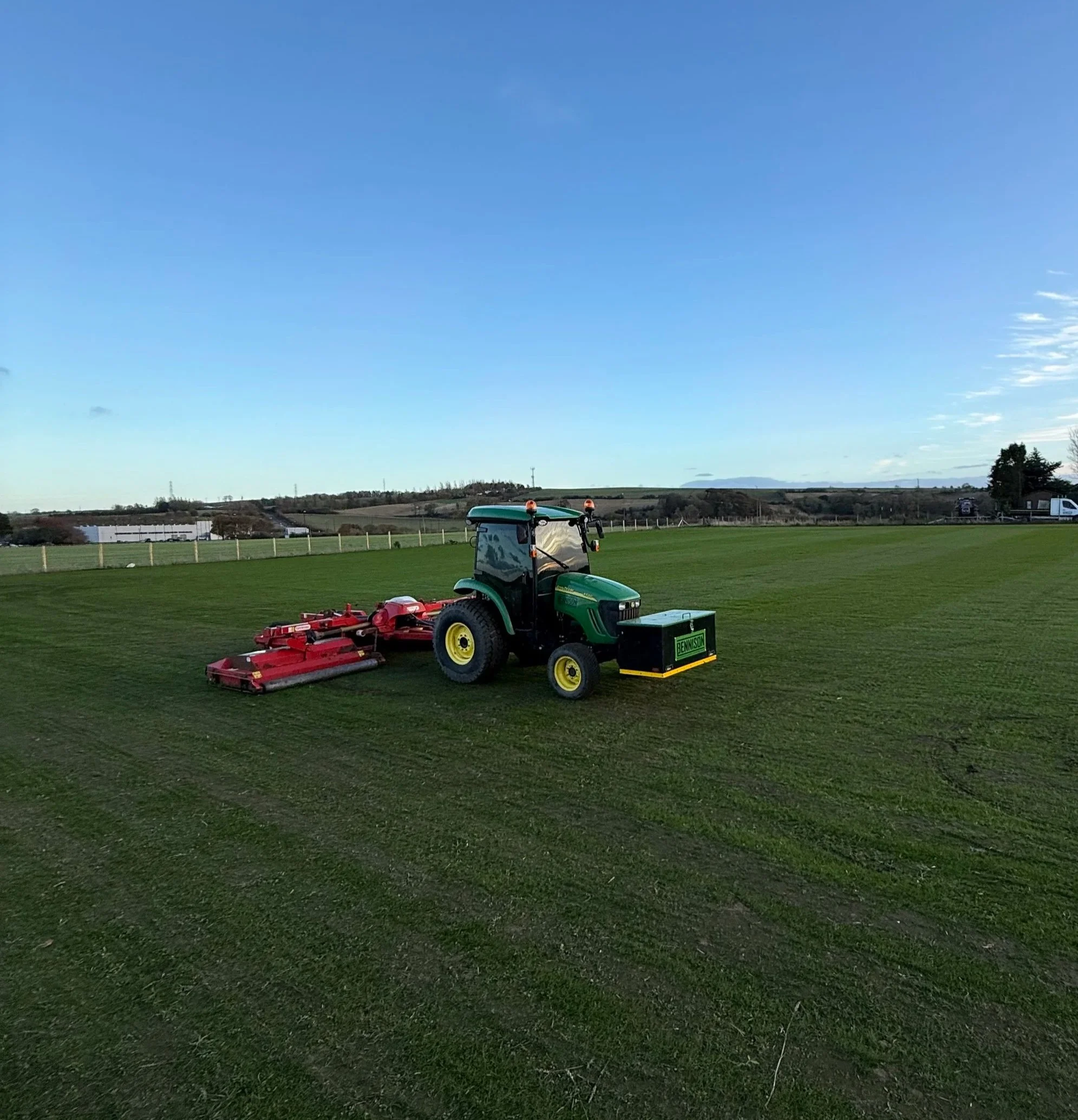 Green and black tractor on a grassy field with a red mower attachment. Clear blue sky and distant hills.