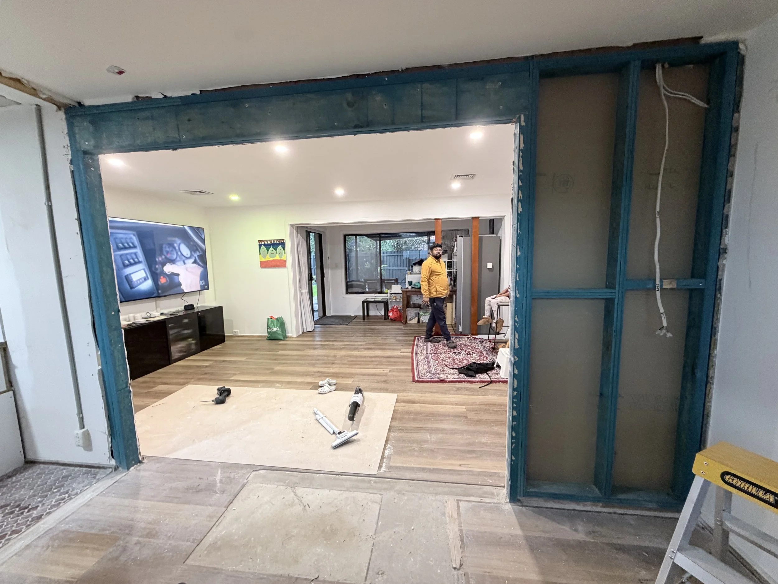 A room undergoing renovation with exposed wooden framing, construction tools on the floor, and a man standing inside.