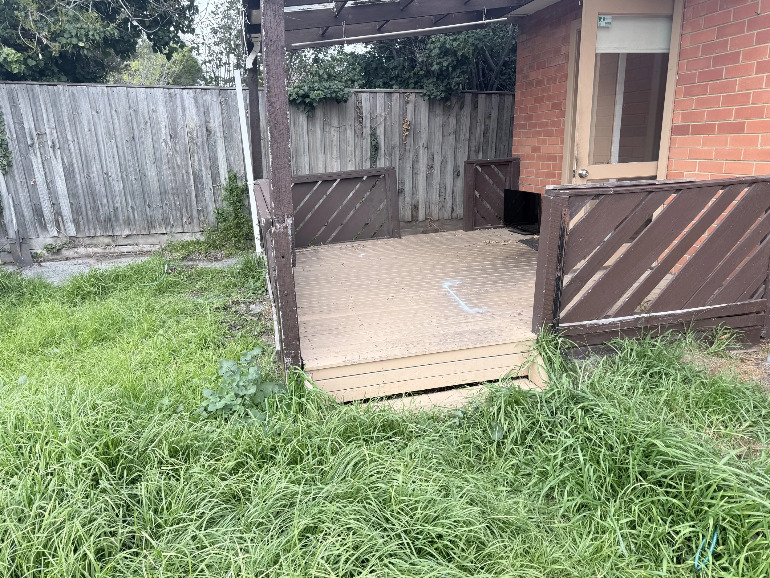Backyard patio with wooden fencing, plants, and a part of a house with a glass door and brick wall. The patio has a wooden deck and surrounding green grass.