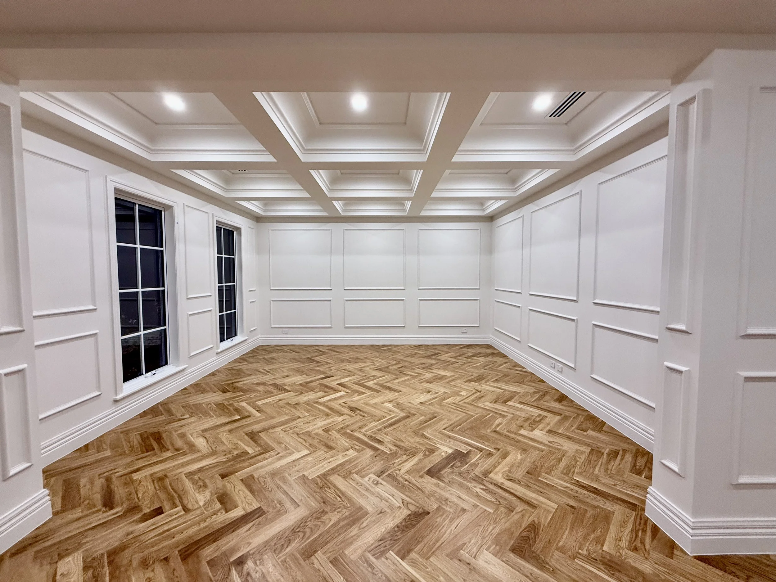 Empty white living room with decorative wall paneling, hardwood herringbone floor, and large windows.