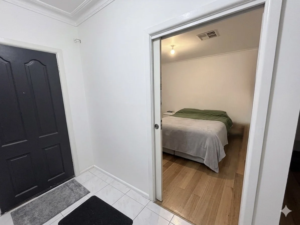 View of a small entryway with a black front door, a gray doormat, and white tiled flooring leading inside a bedroom with a wooden floor, a bed with green and white bedding, a small nightstand, and a ceiling light.