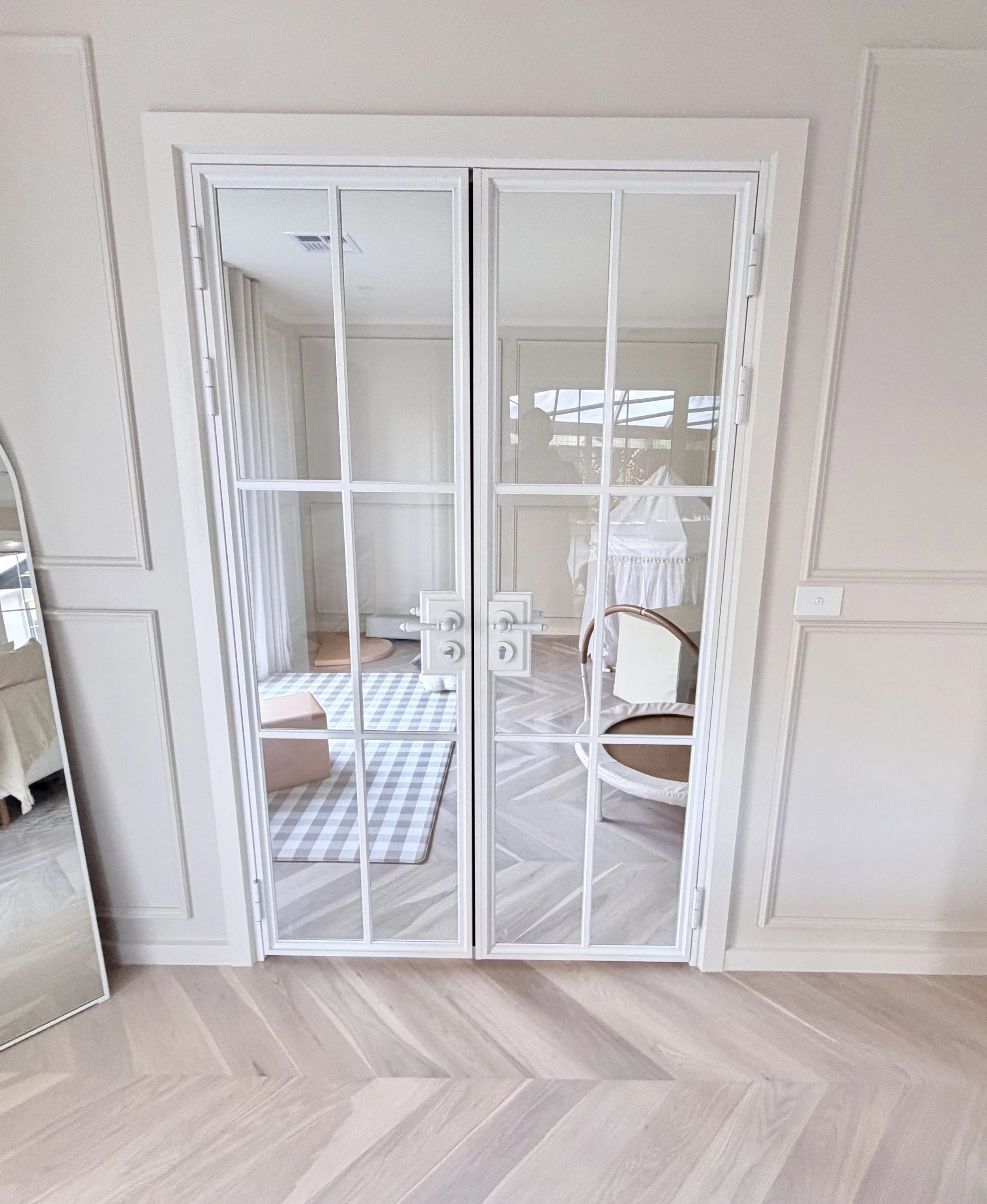 A white double glass door with a white frame and grid design, reflecting a room with windows and furniture inside the house.