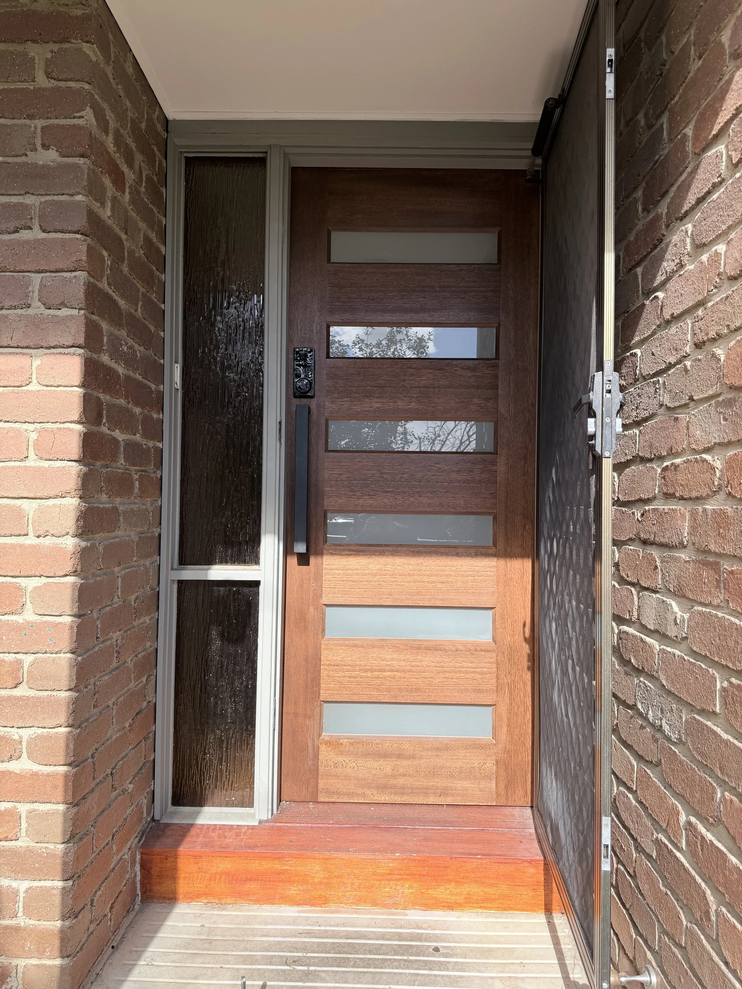 Front door with wooden panels and glass inserts, metal handle, next to a glass sidelights, brick walls, and a metal security screen door on the left.