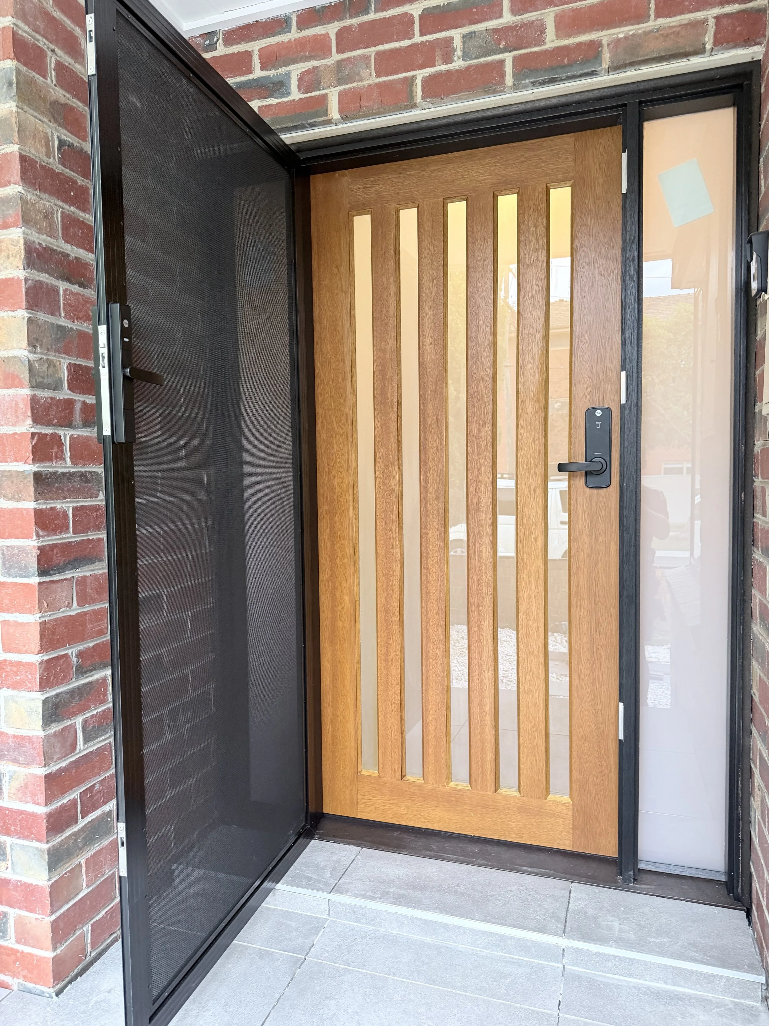A front door with a wooden panel design, embedded within a brick wall, with a screen door open to the left side.