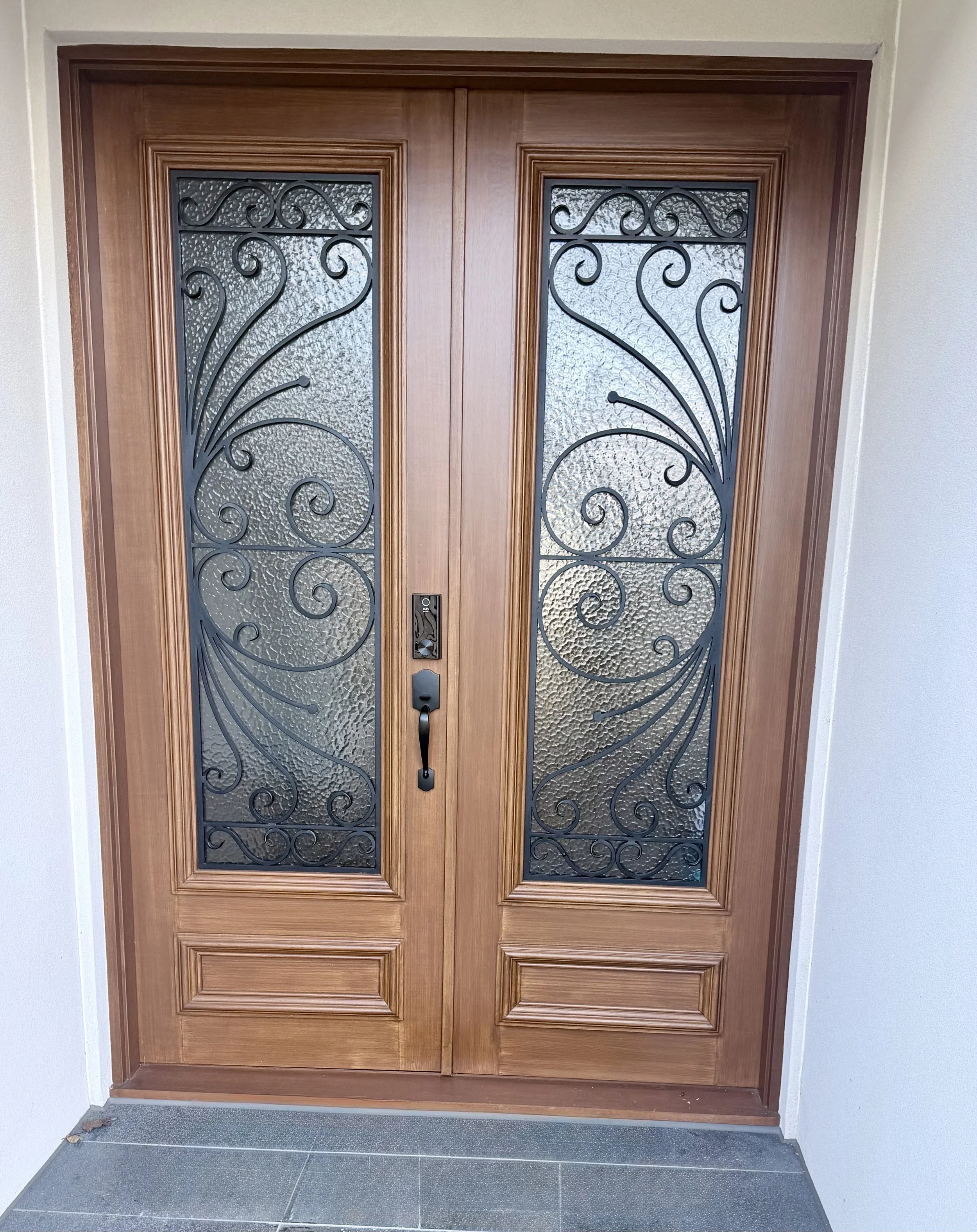 Double wooden front door with decorative black ironwork and textured glass panels, set in a wooden frame, with a handle and lock on the right door. The floor in front of the door has dark tiles, and the wall surrounding the door is light-colored.