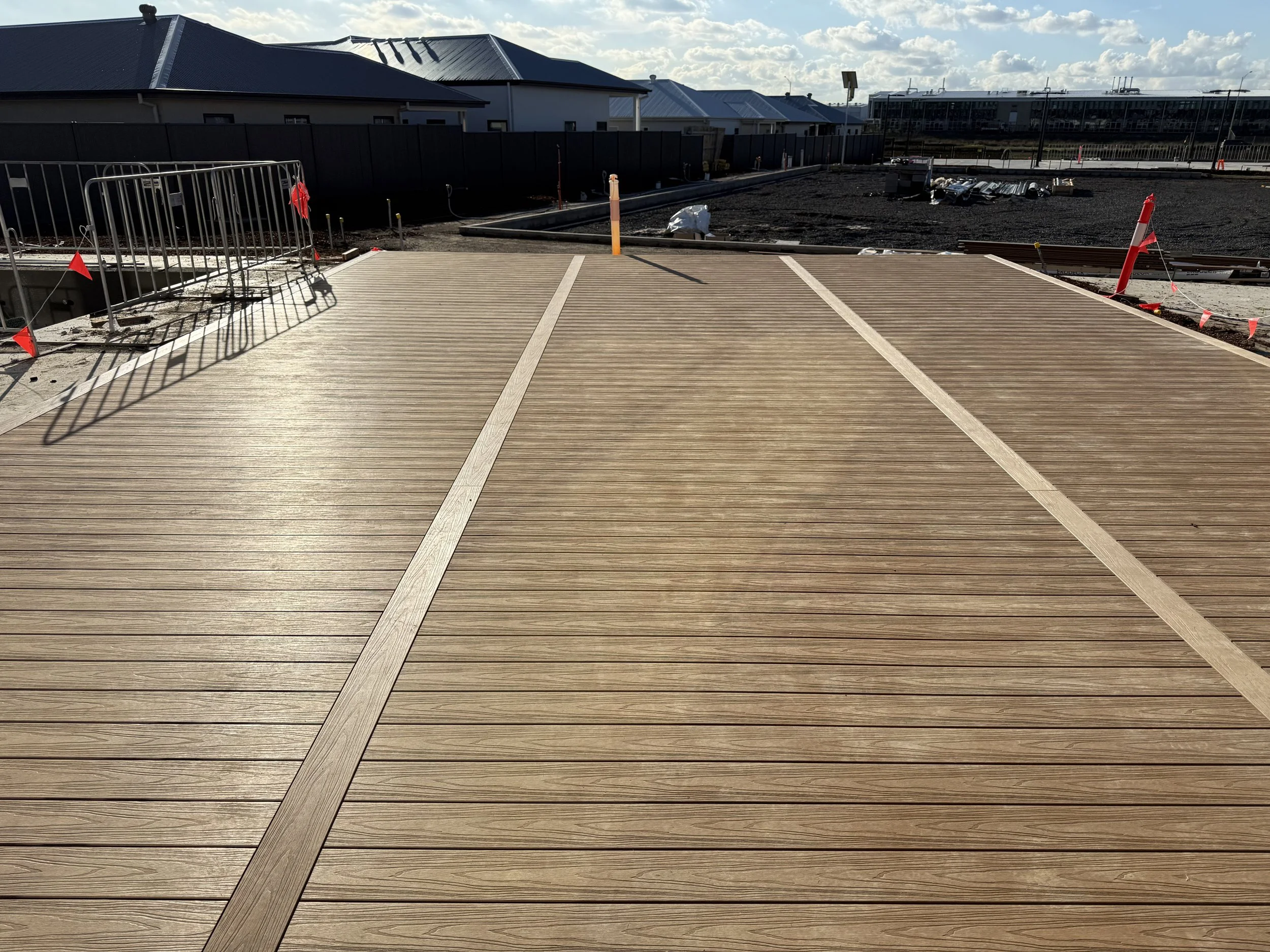 Construction site with a newly installed wooden deck in the foreground, orange traffic cones and a metal barrier to the left, ongoing construction work with building materials scattered on dark soil, houses with metal roofs in the background, a partl
