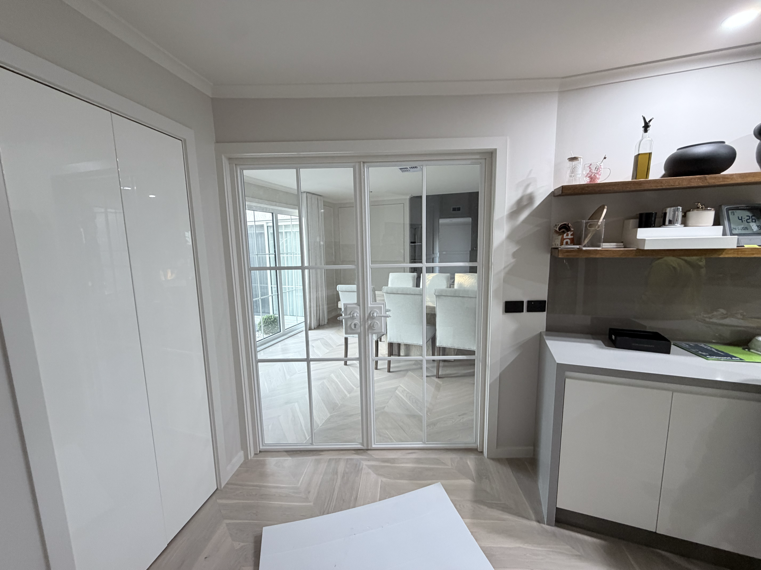 A view through a sliding glass door into a dining area with chairs and a table, with glass panels and a window in the background. Part of a kitchen with shelves holding decorative objects is visible on the right.