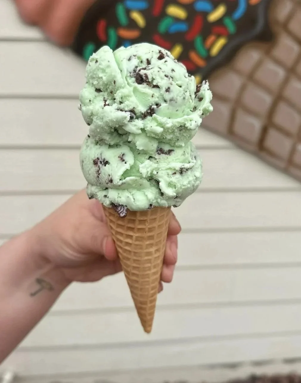 A hand holding a mint chocolate chip ice cream cone with two scoops in a waffle cone, against a background of horizontal white siding and a blurred person wearing a black shirt with colorful sprinkles design.