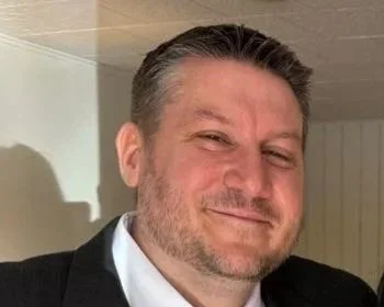 Close-up of a smiling man with short, dark hair and a beard, wearing a black suit and white shirt.