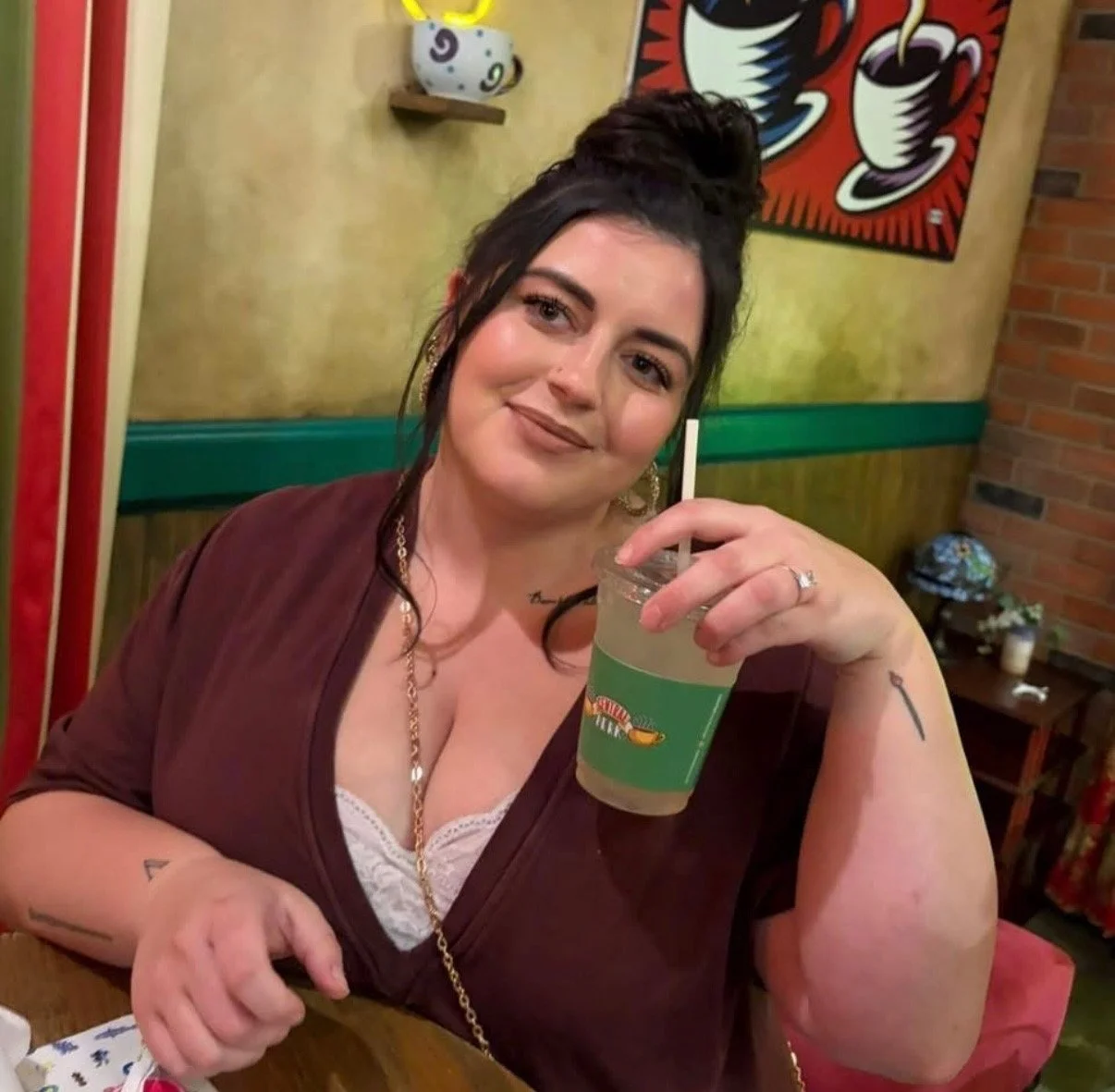 A woman with dark hair in a bun, wearing a maroon top and gold jewelry, sitting at a table in a restaurant, holding a large drink with a straw. Behind her, colorful paintings and decorations are visible.