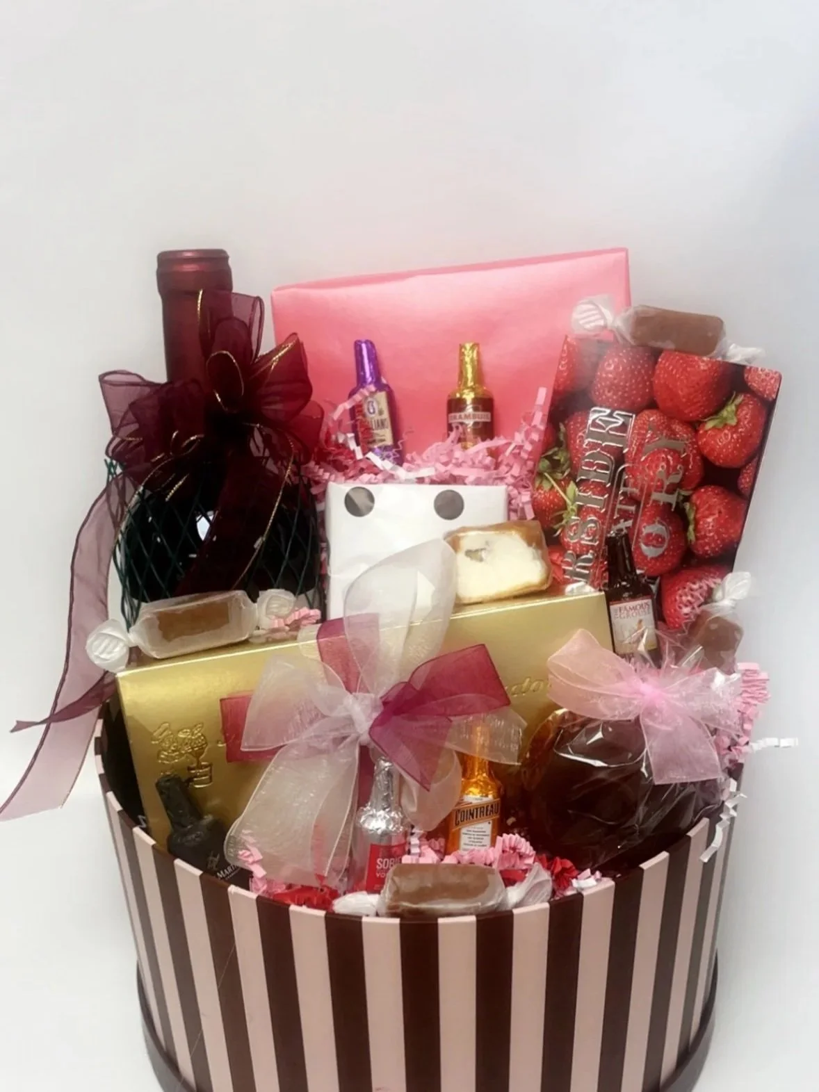 Gift basket with strawberries, bottle of wine, chocolates, and bath products, decorated with pink and brown ribbons.