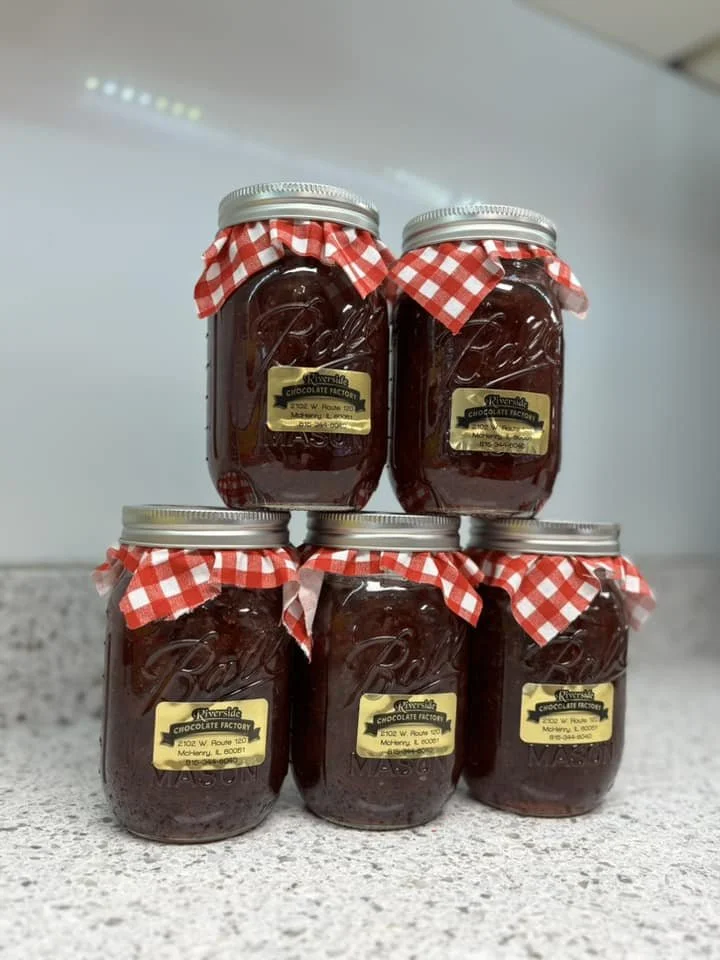 Six mason jars filled with homemade jam, each topped with red and white checkered fabric secured with a silver lid, arranged in a pyramid on a kitchen countertop.