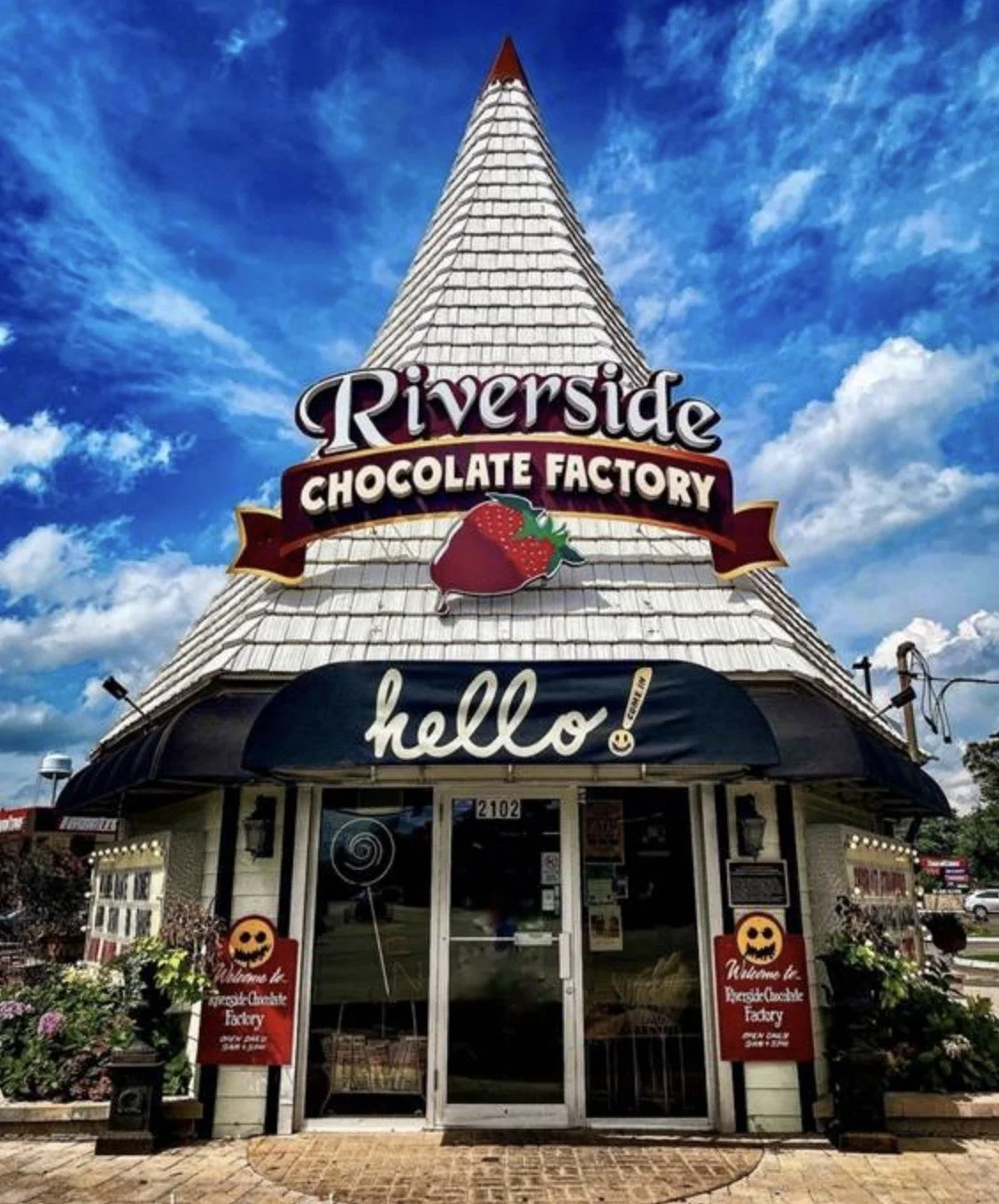 Exterior of Riverside Chocolate Factory with a triangular white shingle roof, a black awning with 'hello' written on it, and a strawberry-shaped sign above. Decorative flowers are outside the entrance, and a clear sky with clouds is in the background.