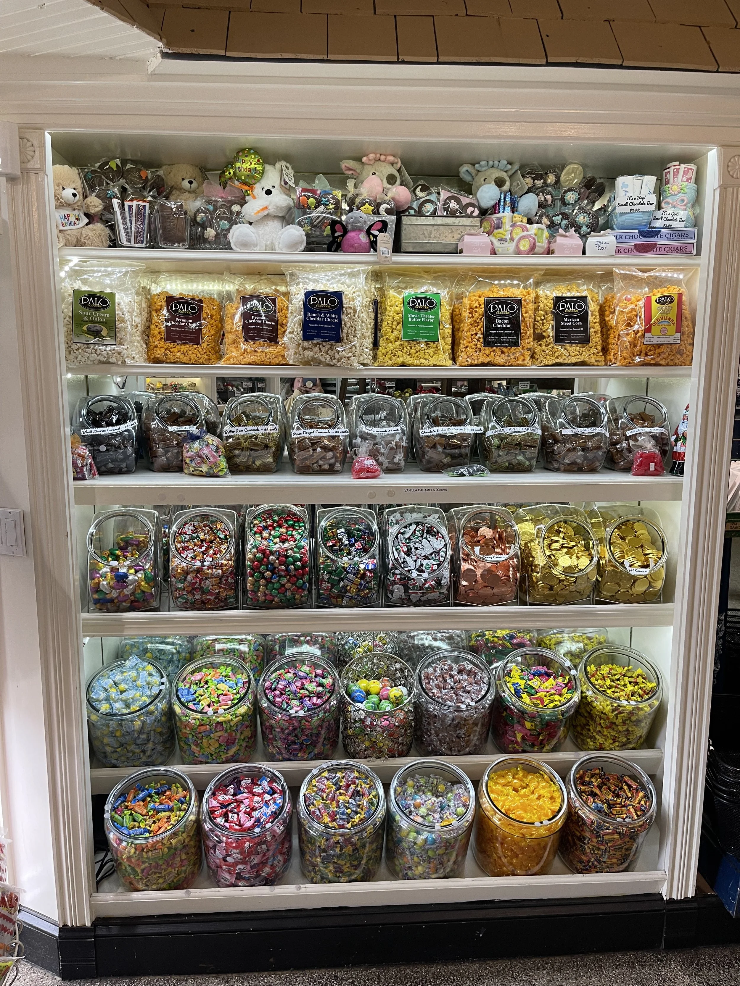 Candy display shelf with multiple clear jars filled with colorful candies and chocolates, and snack bags of popcorn on the top shelf.