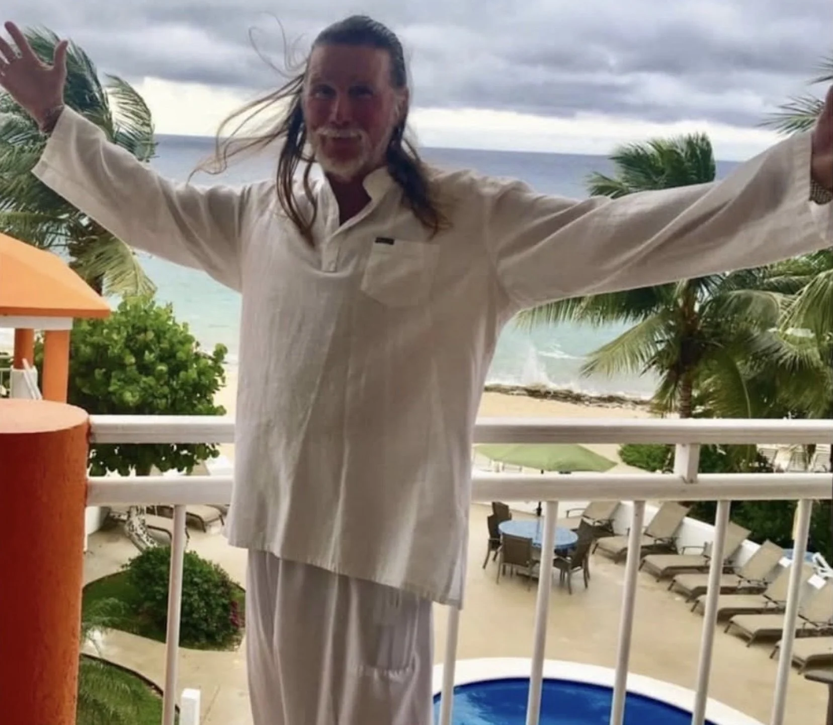 A man with long hair and a beard standing on a balcony with arms outstretched, overlooking a tropical beach with palm trees and the ocean, under cloudy skies.