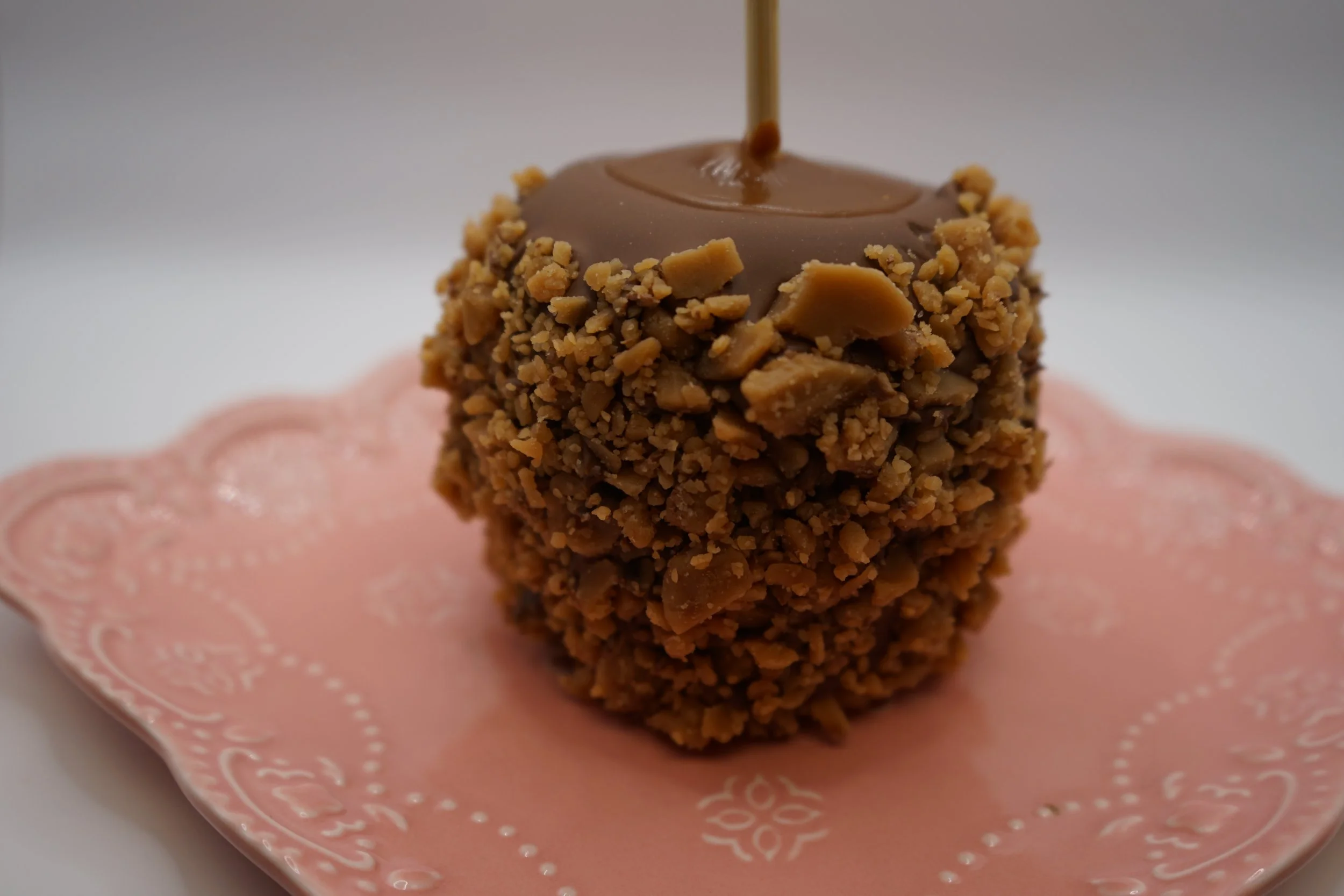 A dessert with a ball coated in crushed nuts, topped with a chocolate drizzle, on a pink decorative plate.