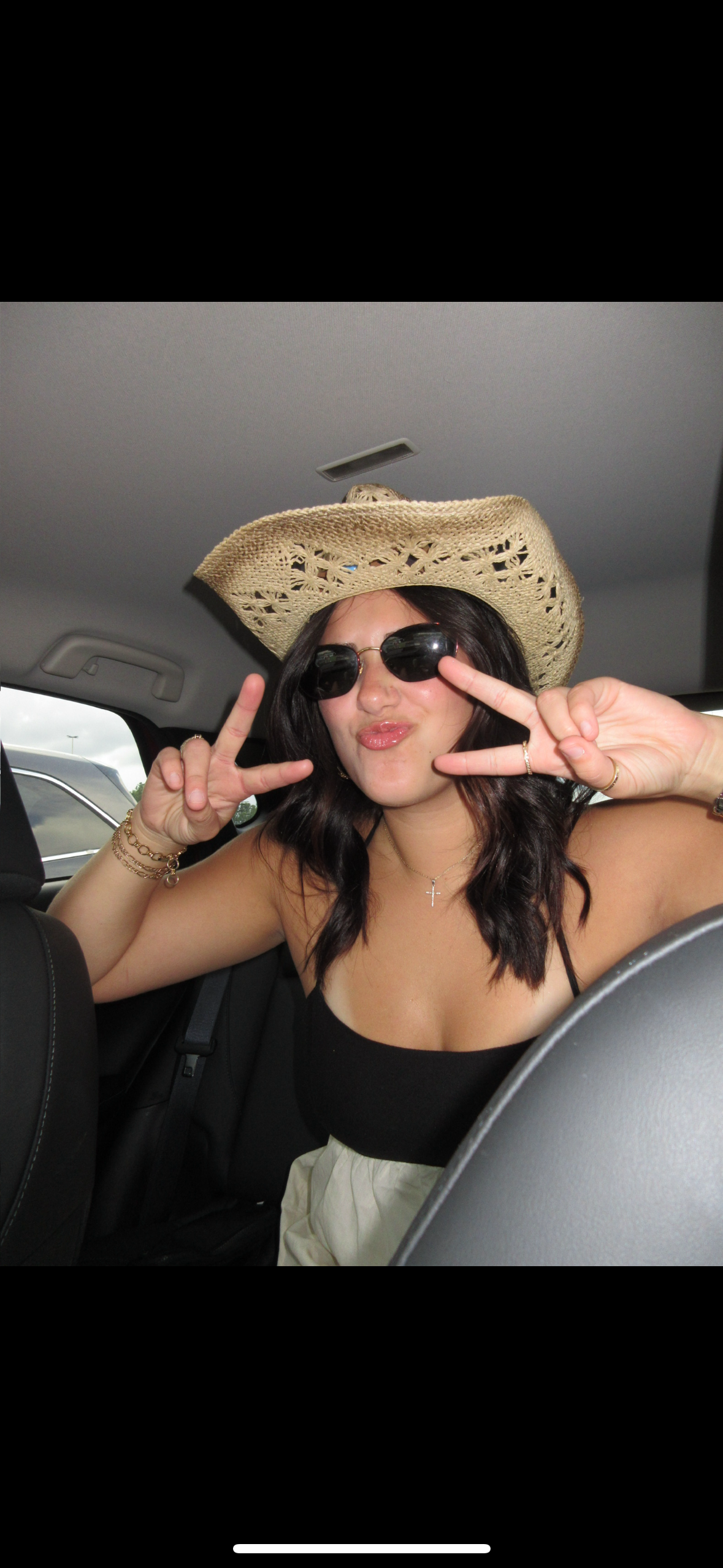 A woman wearing a straw hat and sunglasses, making a peace sign with both hands inside a car.