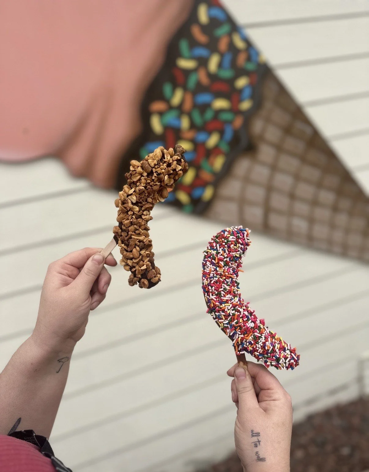 Two hands holding banana-shaped ice cream bars decorated with sprinkles. In the background, part of a brick building with colorful frosting and sprinkles is visible.
