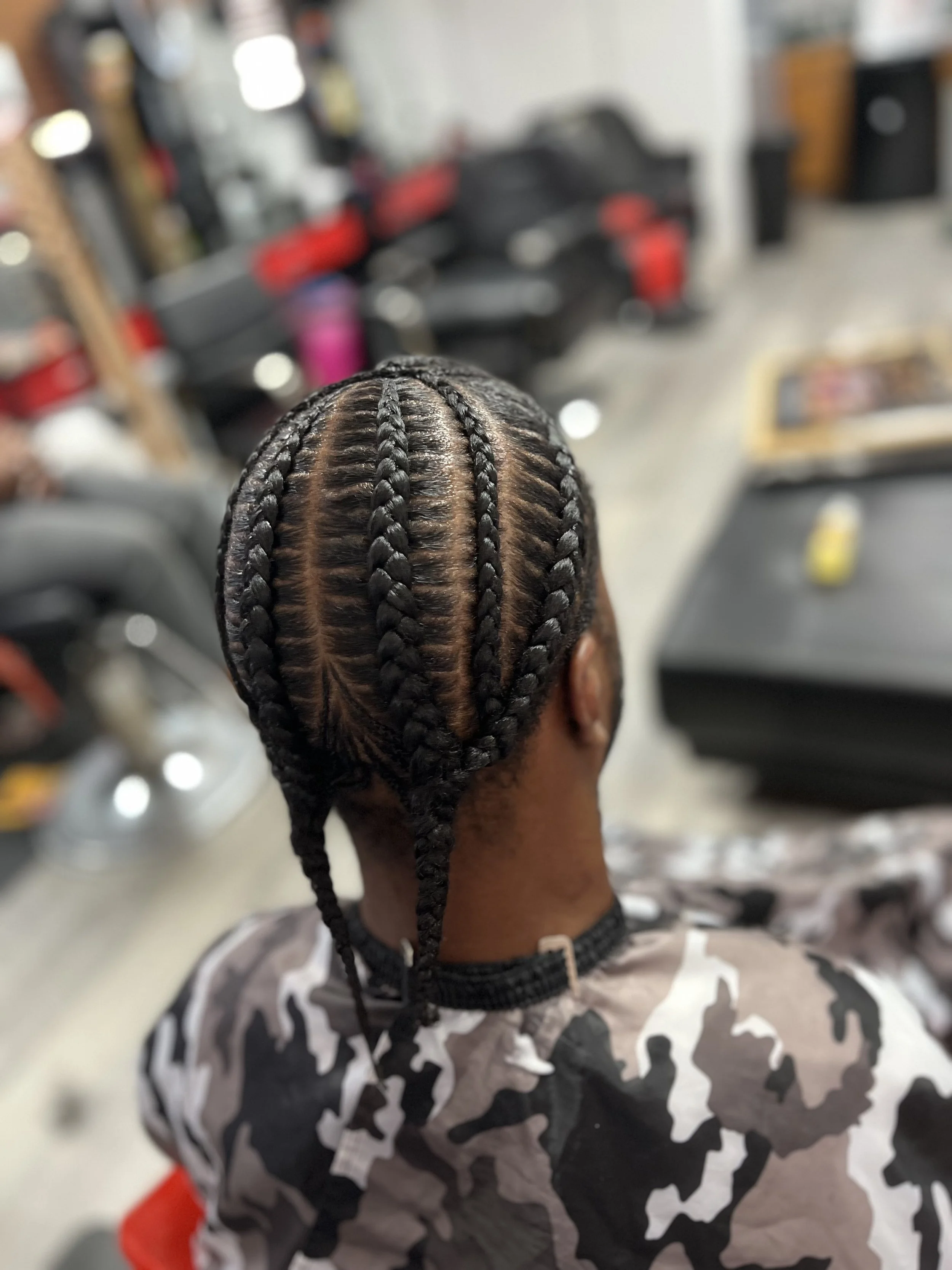 A person with neatly braided hair sitting in a barbershop or salon, with blurred background showing hair styling equipment and products.
