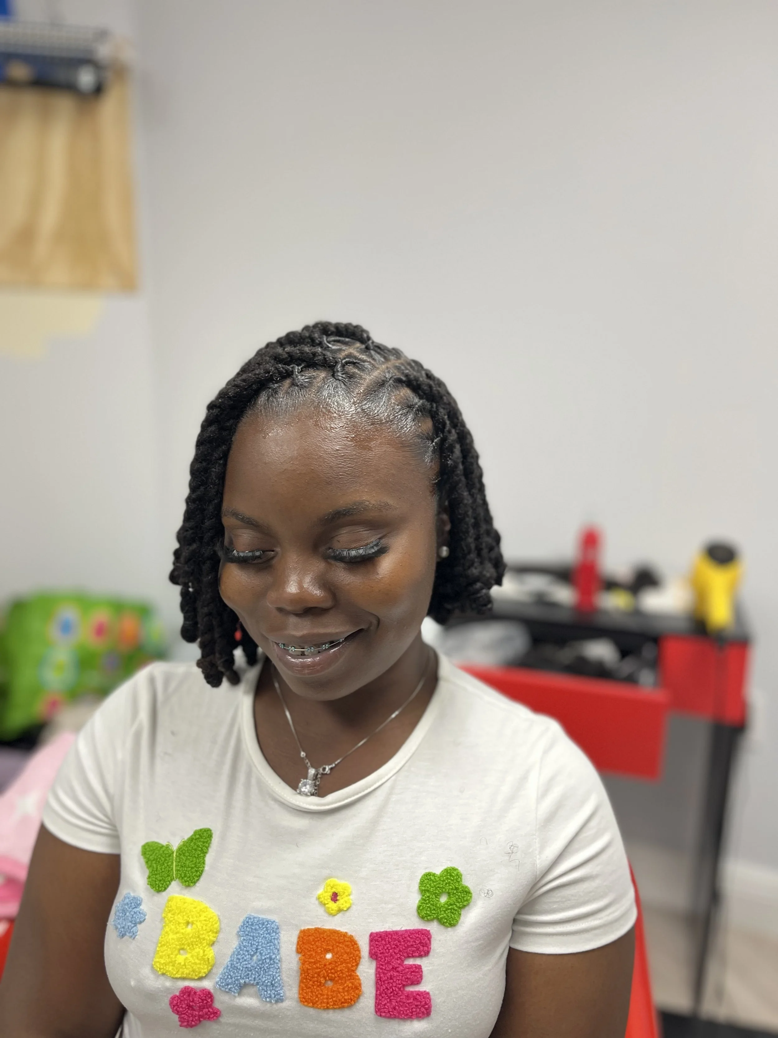 A woman with dark braided hair, wearing a white t-shirt with colorful felt letters spelling 'BABE' and felt flowers and butterflies, smiling and looking downward, in a room with a red toolbox and supplies in the background.