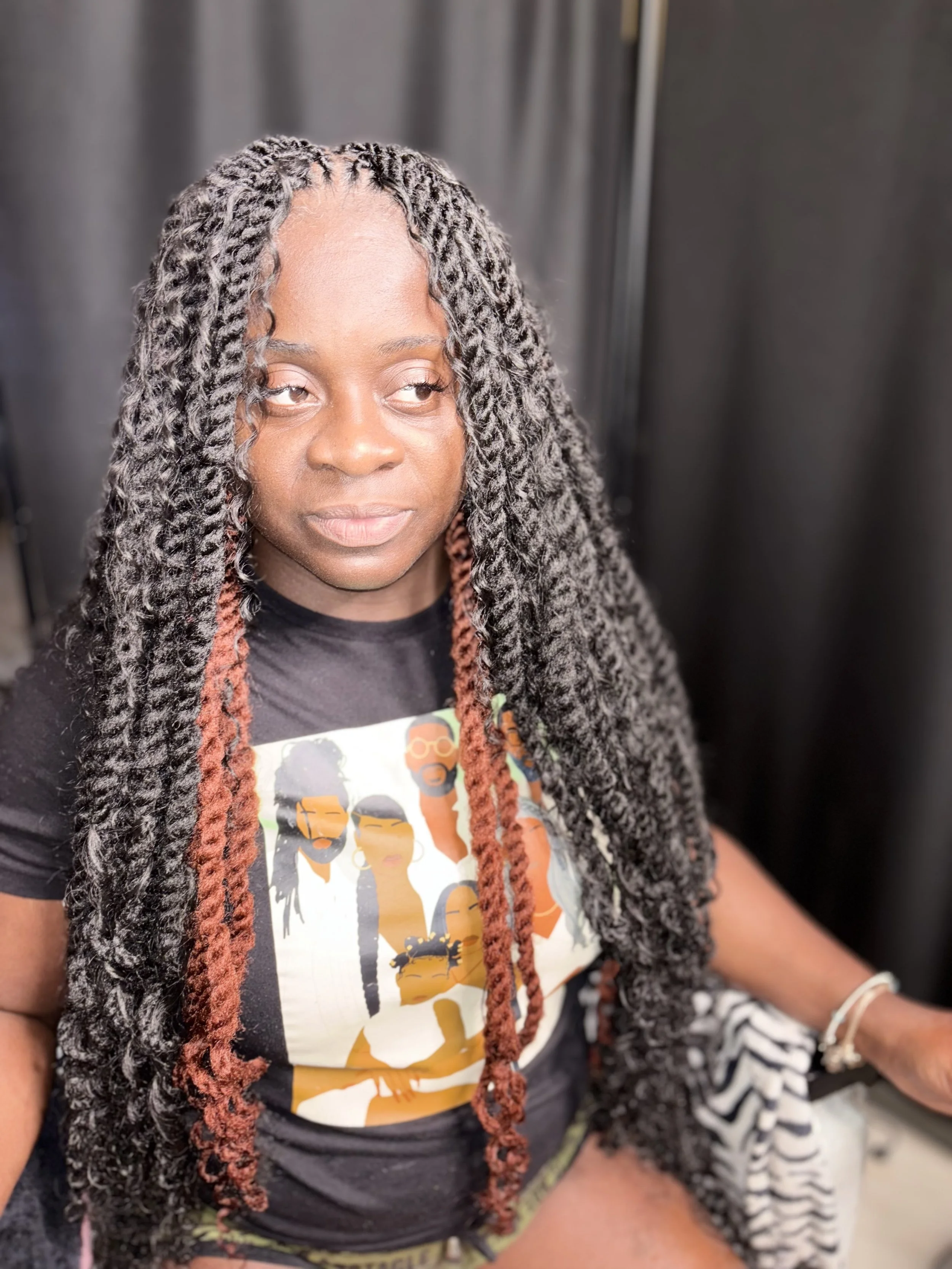 A woman with long, twisted hair featuring dark and reddish-brown colors, sitting in front of a black curtain, wearing a black graphic t-shirt with illustrations of women and sunglasses.