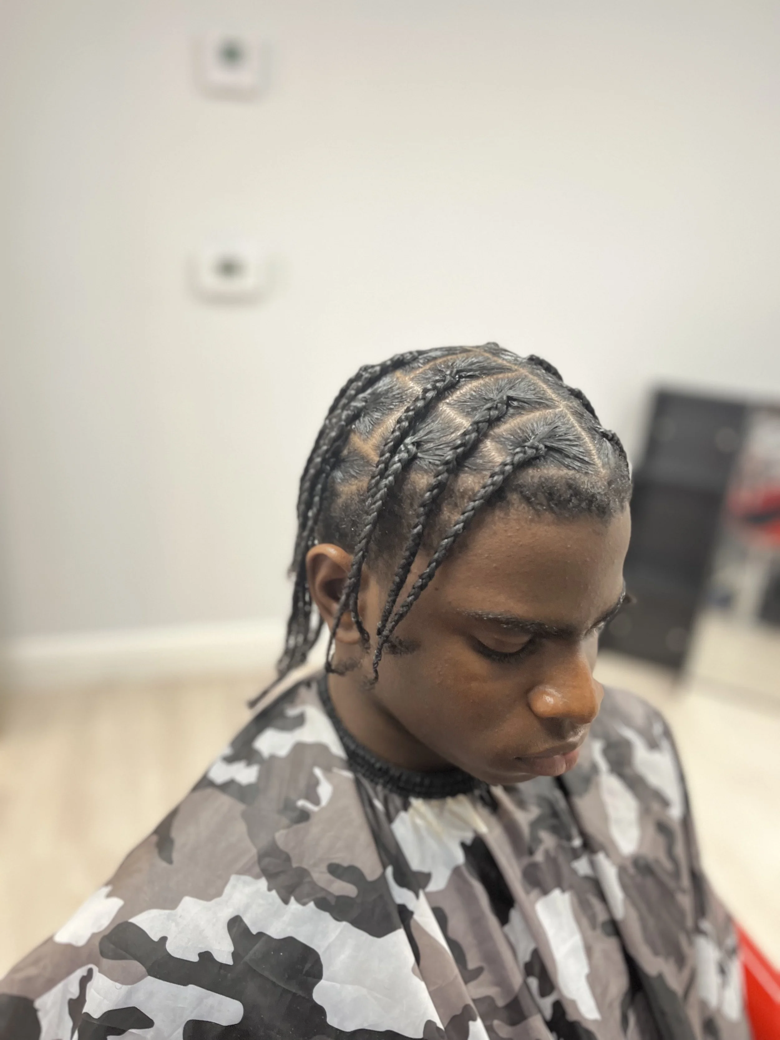 Young man with braided hair getting a haircut in a barbershop, wearing a camo-patterned cape.