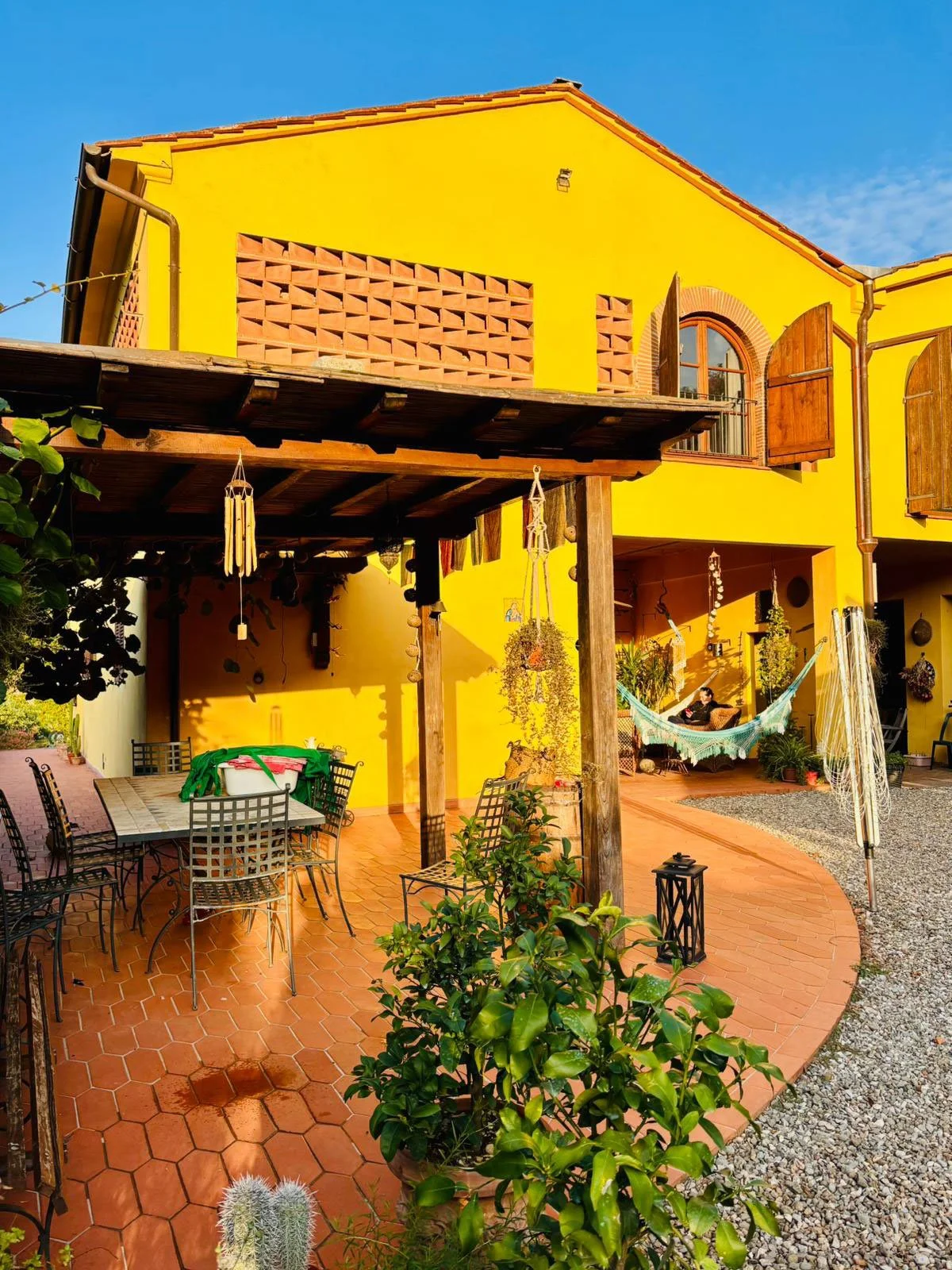 Yellow house with wooden window shutters, outdoor patio with table and chairs, hanging plants, hammock, and green plants, under a clear blue sky.