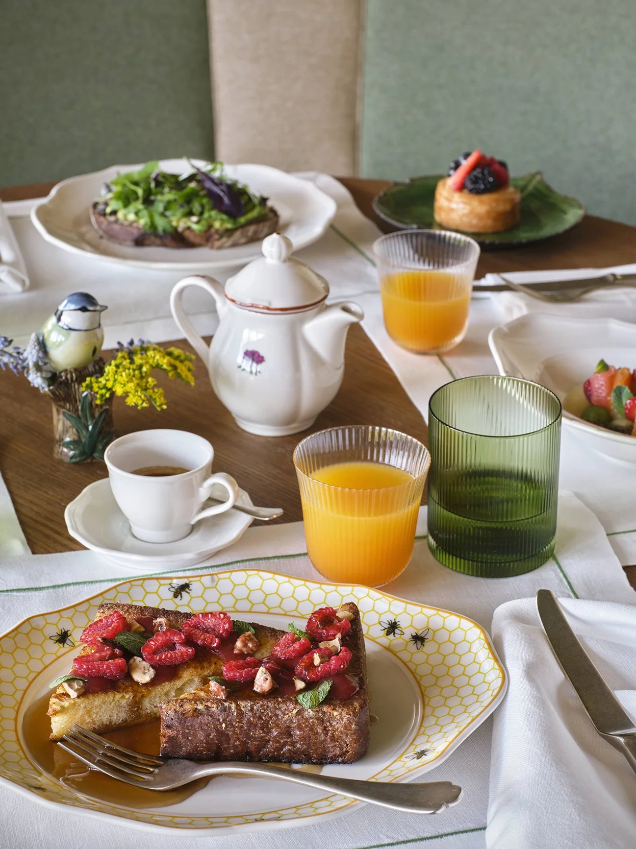 A table set with breakfast items including a slice of cake with strawberries and nuts, a cup of coffee, two glasses of orange juice, a teapot, a green drinking glass, a salad, a piece of toast with avocado and greens, a muffin with berries, and a small bird figurine with flowers.