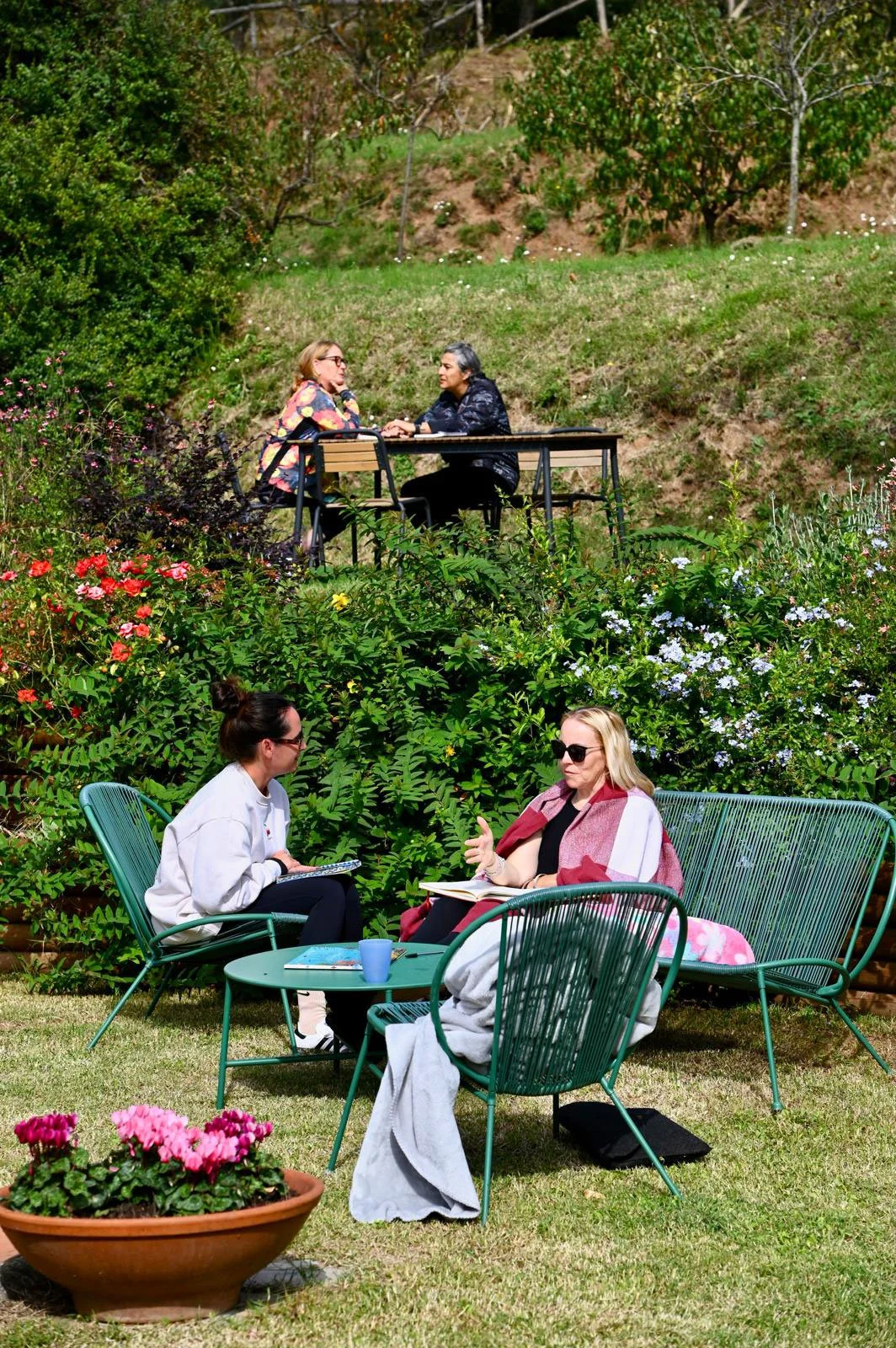 Two women sitting outdoors on a green metal table and chairs, having a conversation on a grassy lawn with a flowerpot in the foreground, and two other women sitting at a table on a small hill in the background, surrounded by lush greenery and flowers.