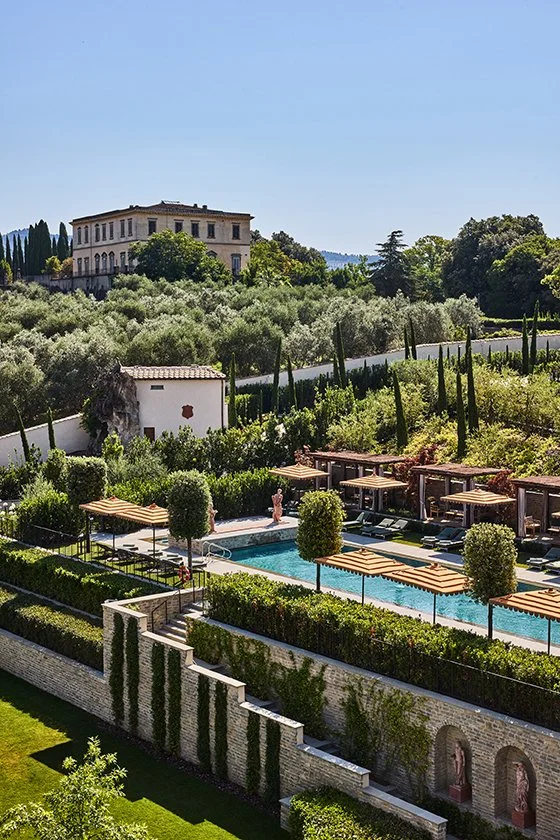 Luxury outdoor swimming pool area surrounded by greenery, umbrellas, and cabanas with a historic building and hills in the background.