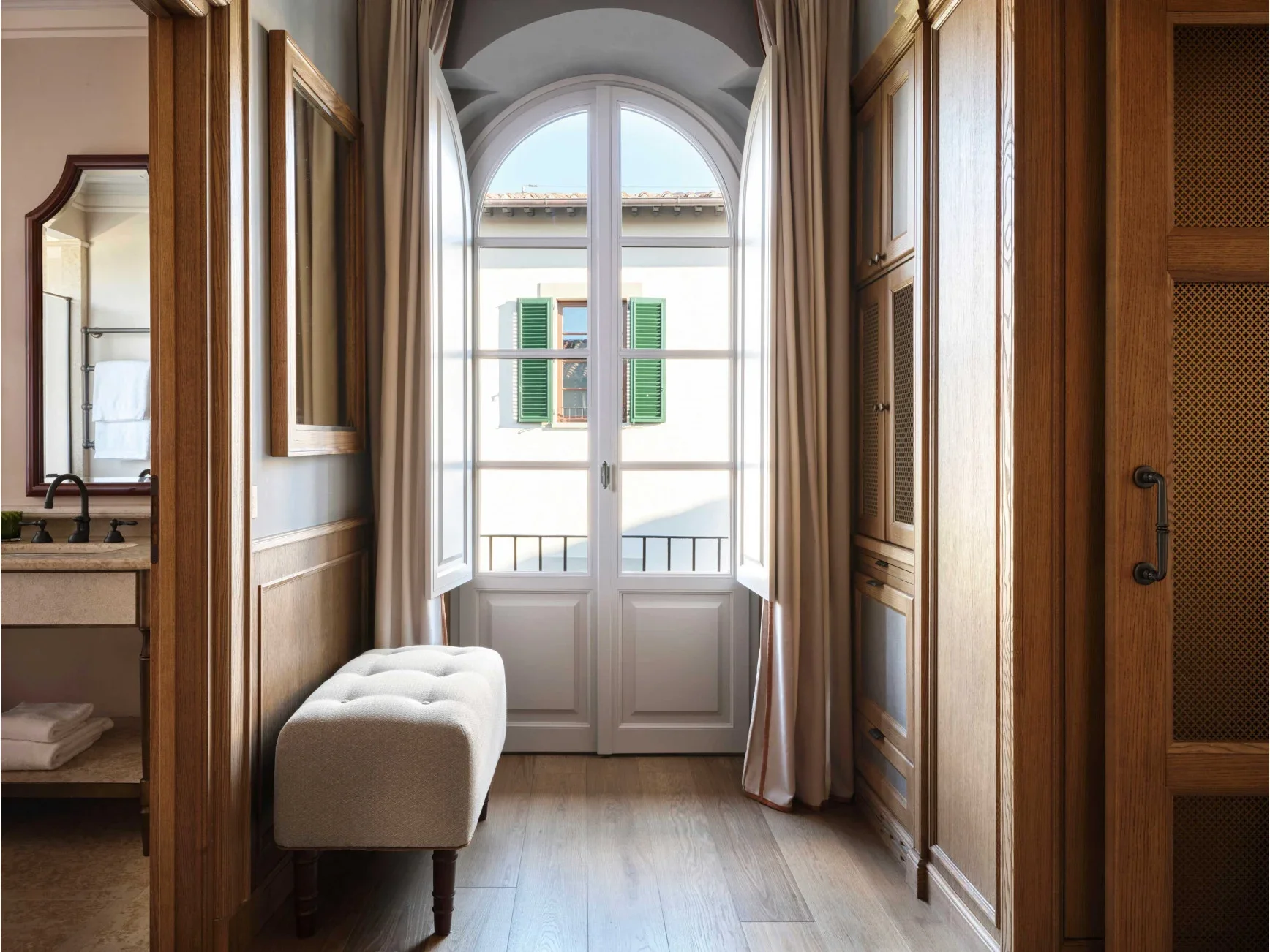 A view of a bright room with a large arched window with open shutters, beige curtains, a beige bench, wooden cabinets, and a glimpse of an adjacent bathroom with towels.