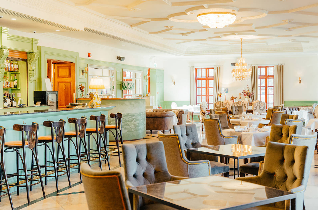 Elegant restaurant or hotel dining area with large windows, upholstered chairs, and a bar with high stools.