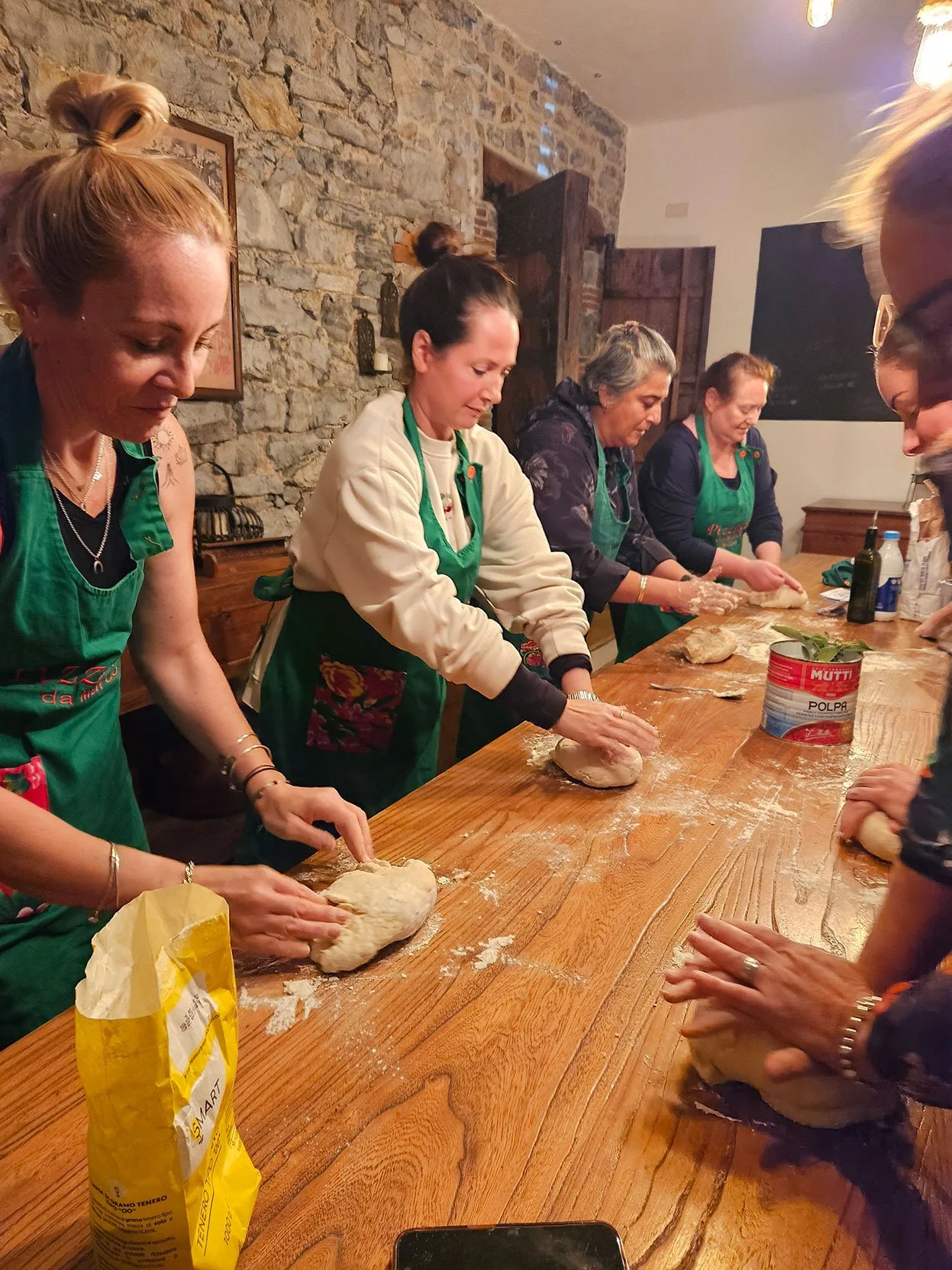 A group of women participating in a dough-making activity around a wooden table in a rustic room with stone walls.