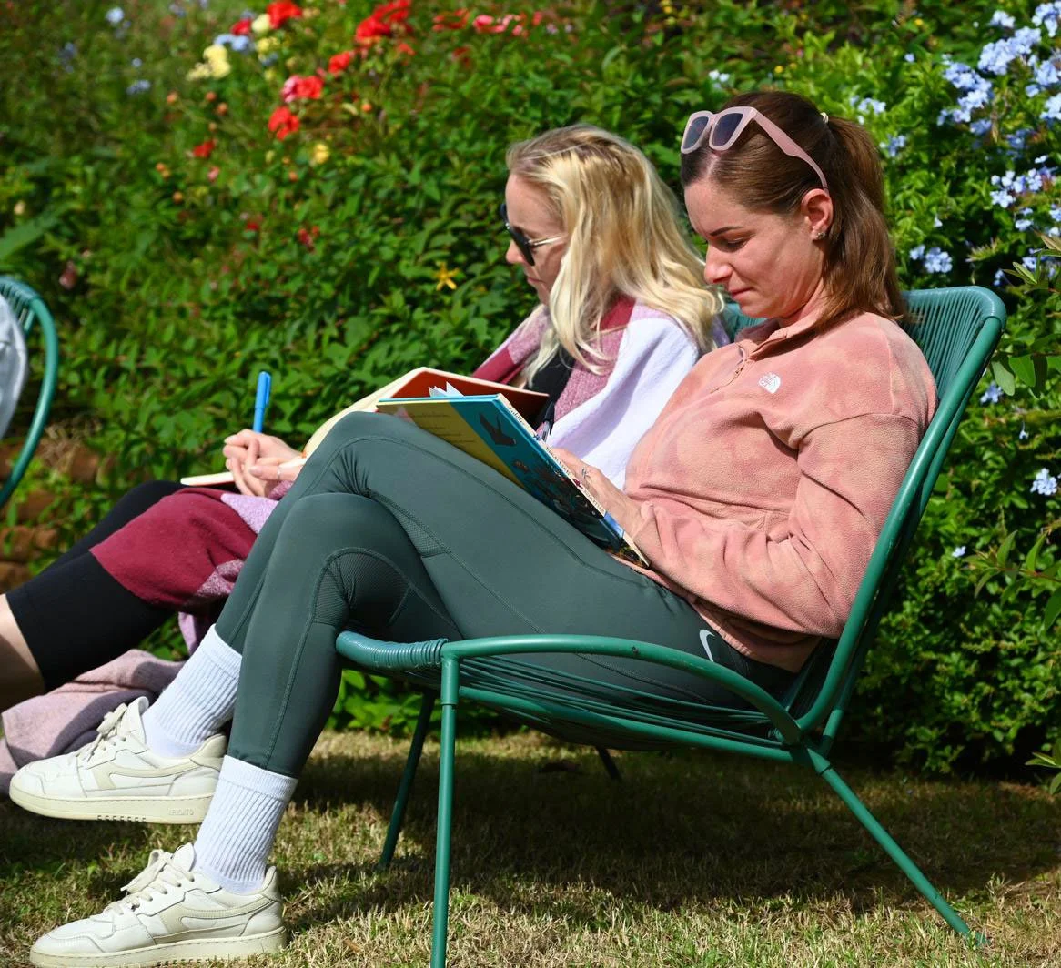 Two women sitting on green metal chairs outdoors, reading and writing in notebooks, with lush green shrubbery and colorful flowers in the background.