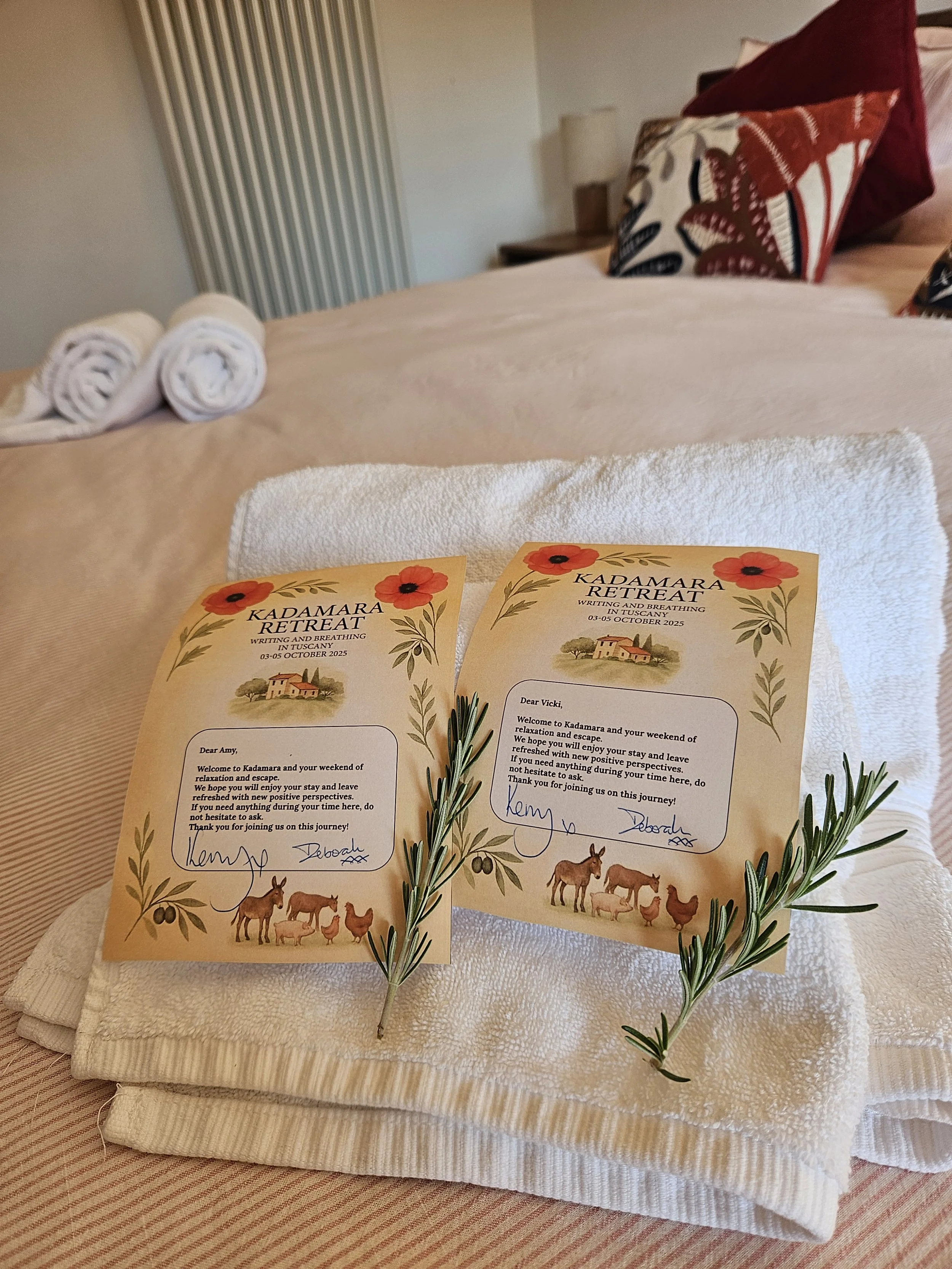 Two welcoming notes from Kadamara Retreat are placed on a folded white towel on a bed. Each note is decorated with illustrations of farm animals, flowers, and greenery, and signed by Kem and Deborah. Fresh rosemary sprigs are placed on the notes.
