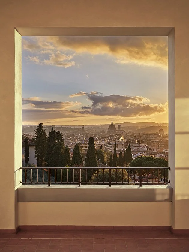 View through a square window showing a cityscape at sunset with hills, trees, and a prominent domed building in the background.