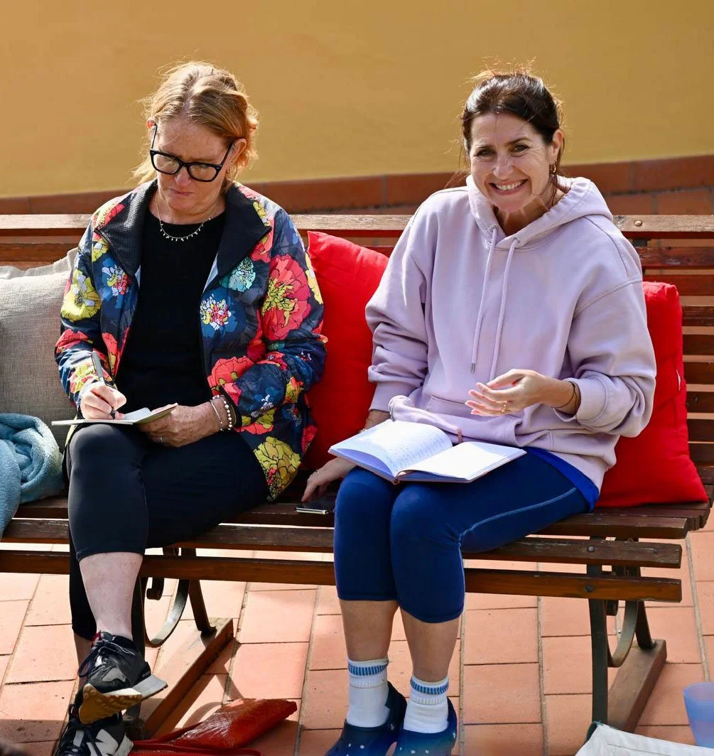 Two women sitting on a wooden bench, one is writing in a notebook and the other is smiling and speaking, with notebooks on their laps.