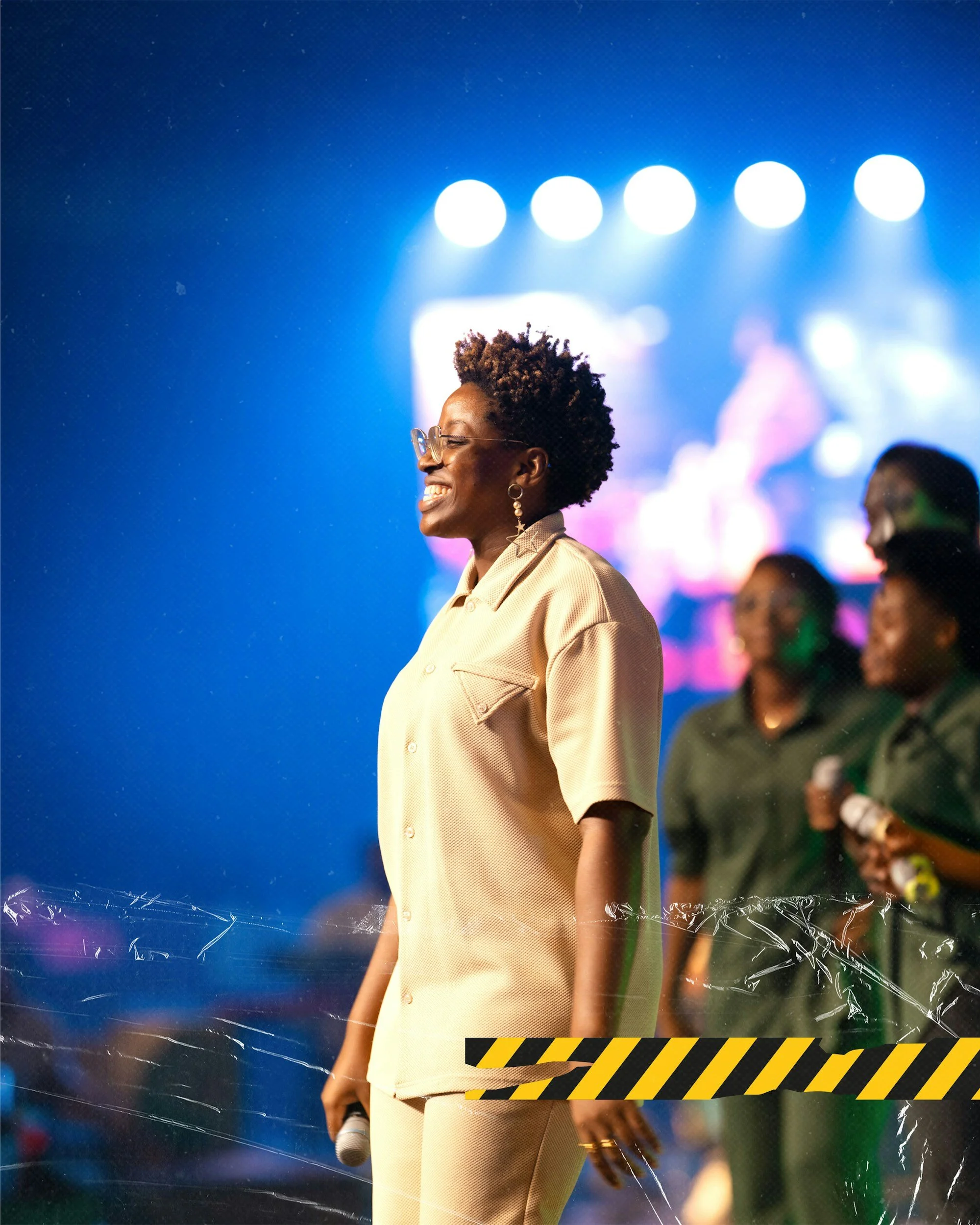 A woman with short curly hair, glasses, and star-shaped earrings smiling on stage at an event, with a blue background and colorful lights, and multiple people in the background.