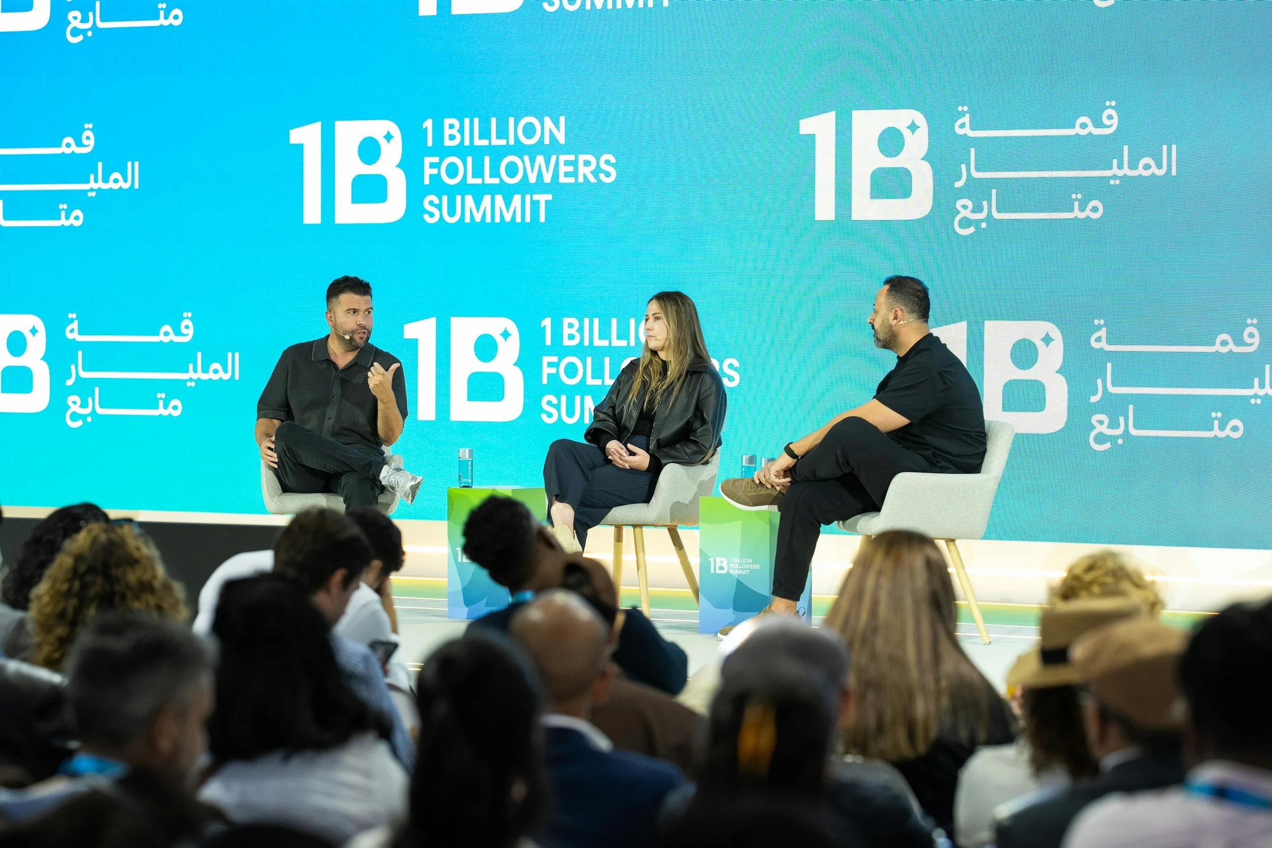 Three people are sitting on stage during a panel discussion at the 1 Billion Followers Summit, with a large blue backdrop displaying the summit's name and logo in English and Arabic. They are engaged in conversation, with audience members visible in the foreground.