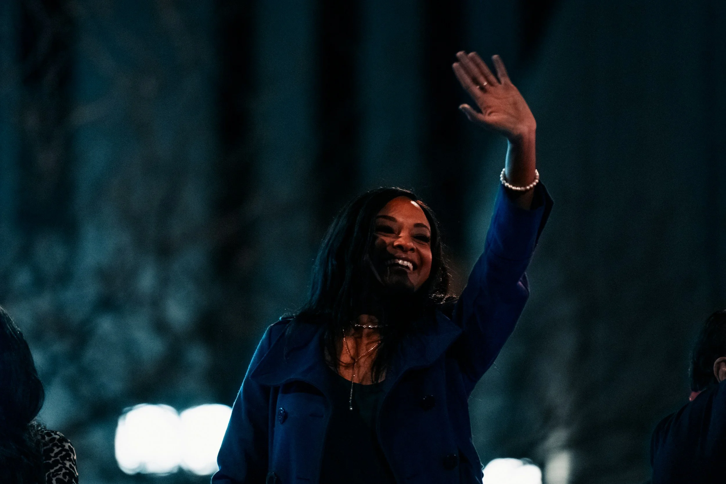 A woman smiling and raising her hand in a dark setting with blue and white lighting.