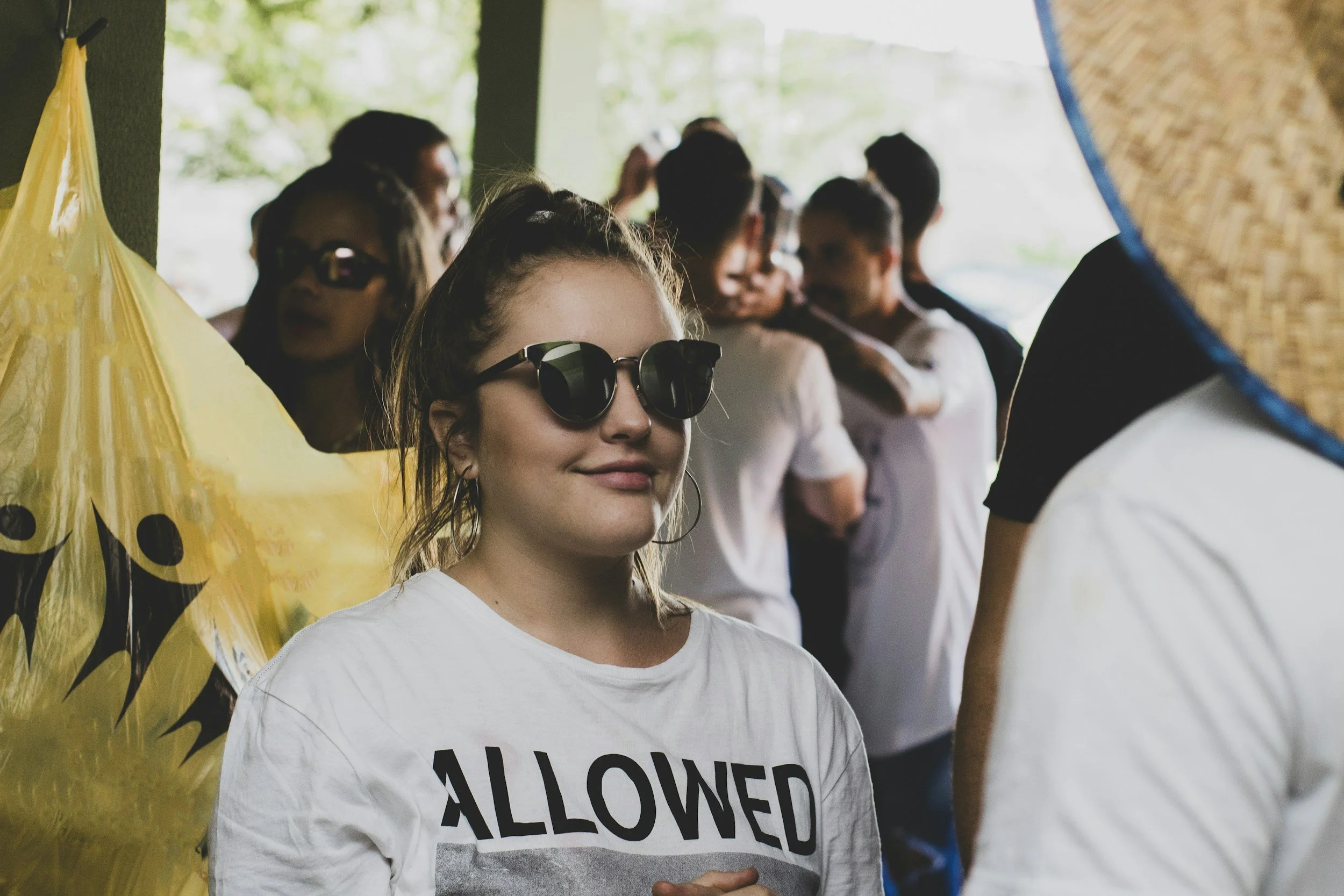 A young woman wearing sunglasses and a white T-shirt with the word 'ALLOWED' printed on it, smiling at an outdoor gathering with other people in the background, some wearing white shirts, and a yellow balloon with smiley face on the left side.