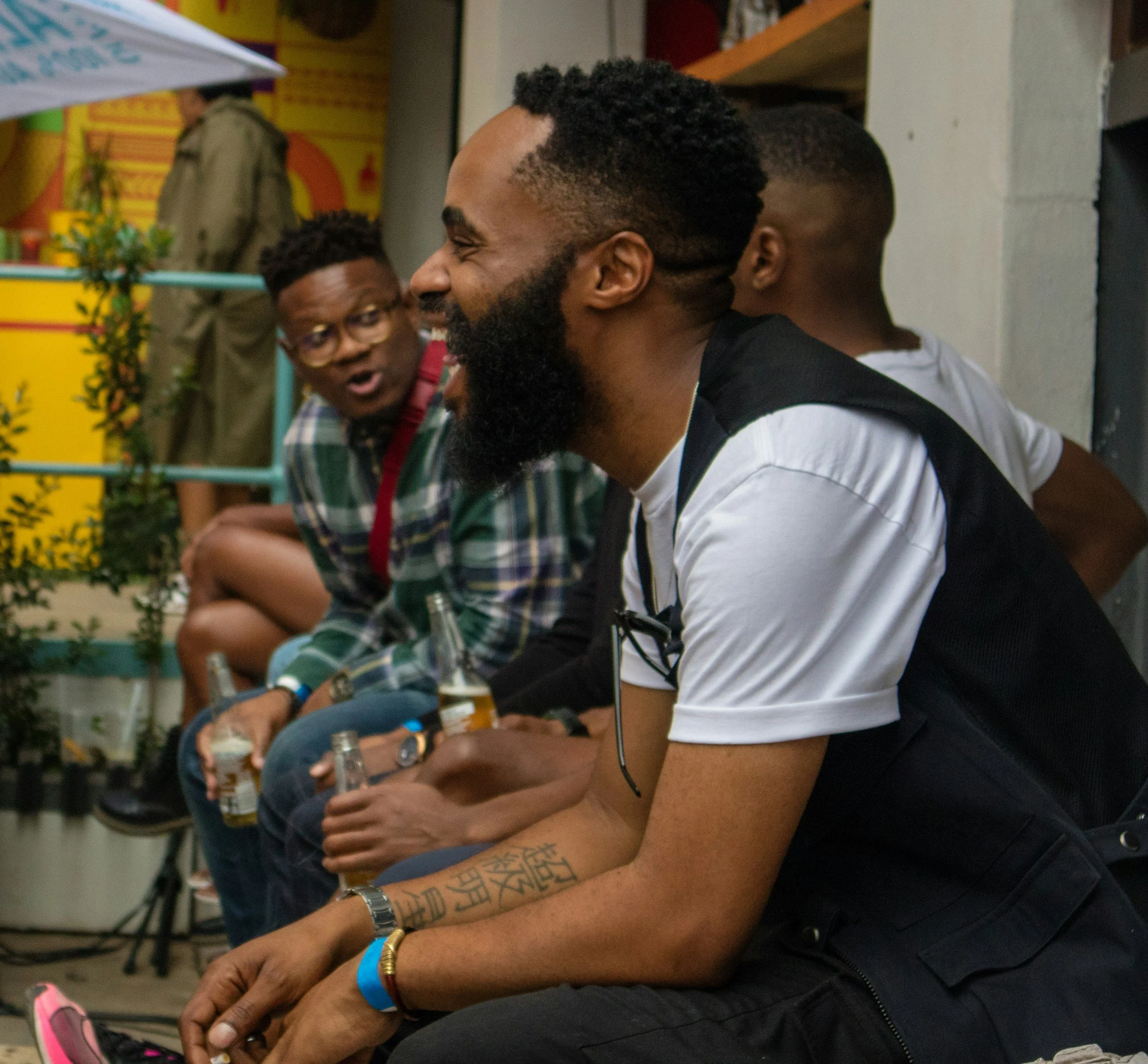 Three men sitting at a table, engaged in conversation. The man in the foreground has a beard, tattoos on his arm, and is wearing a white shirt with black sleeves. The man to his left wears glasses and a plaid shirt. The third man, in the background, is wearing a dark shirt. They are in a casual setting with colorful decorations and plants in the background.
