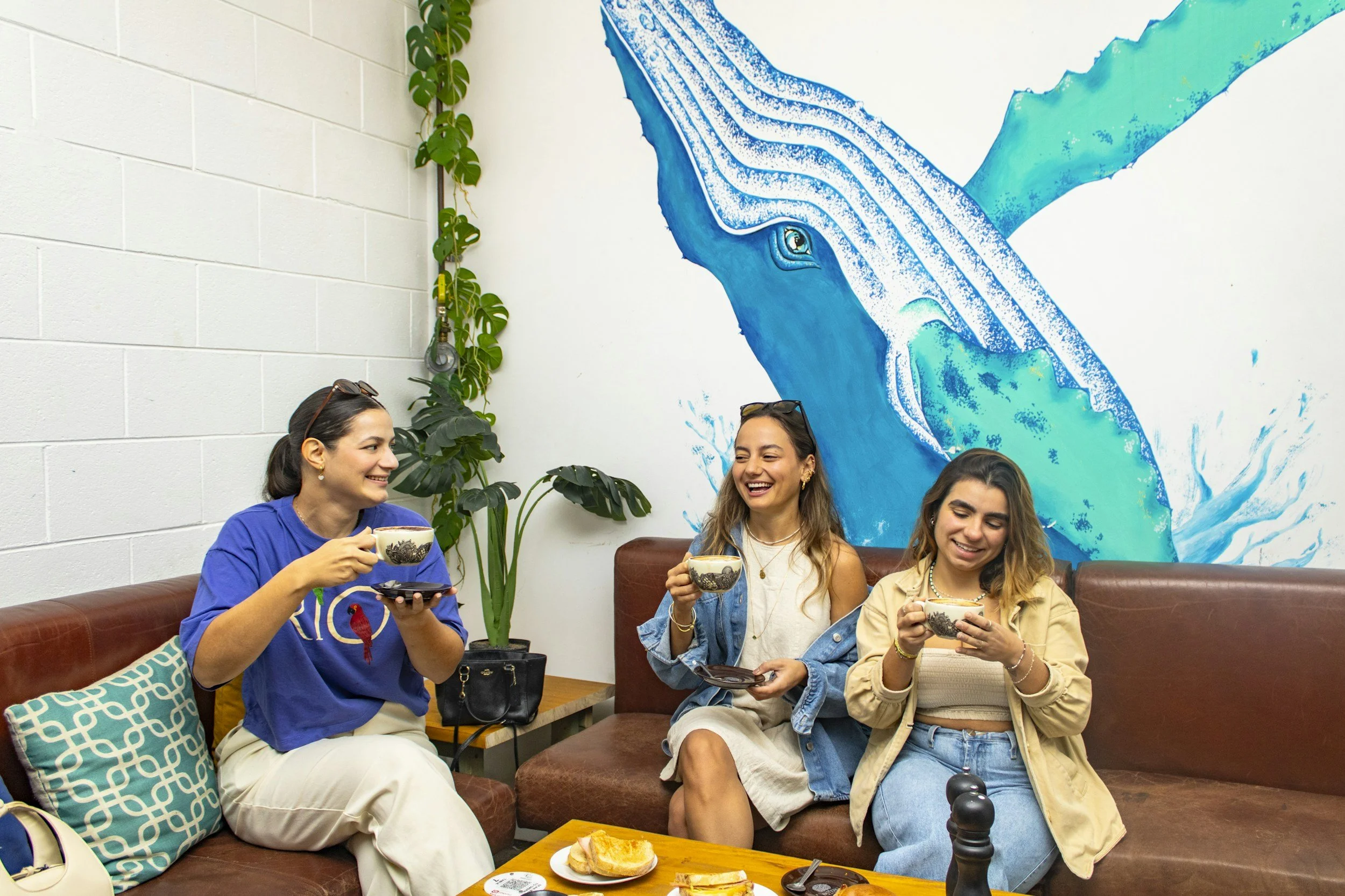Four women sitting on a brown leather sofa, enjoying tea and snacks, smiling and talking. The background features a large blue whale mural on the wall and a green plant climbing the wall.