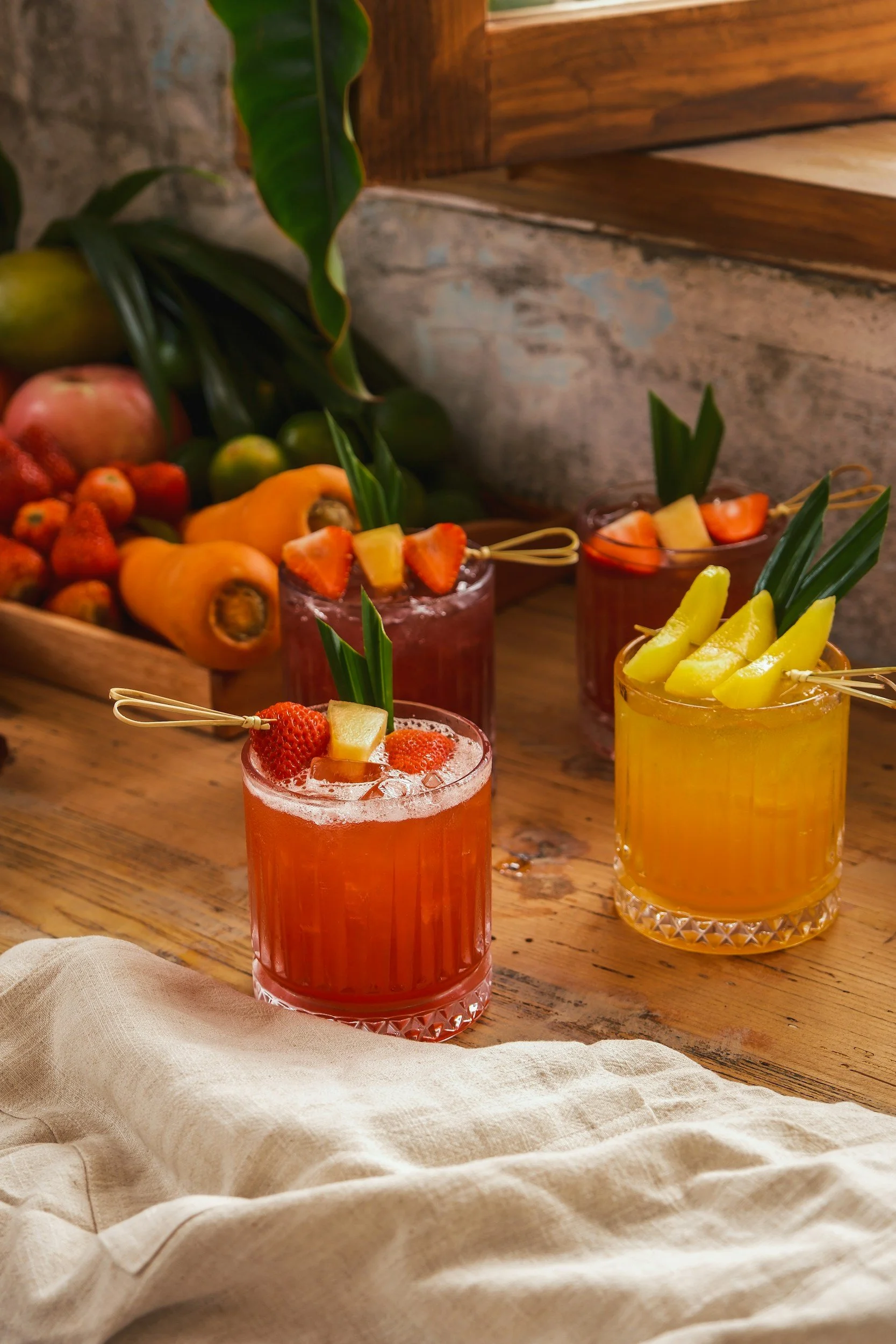 A colorful fruit cocktail with strawberries and pineapple garnishes, served in a textured glass, set on a rustic wooden table with a beige fabric underneath, and surrounded by an assortment of fresh fruits and green leaves.